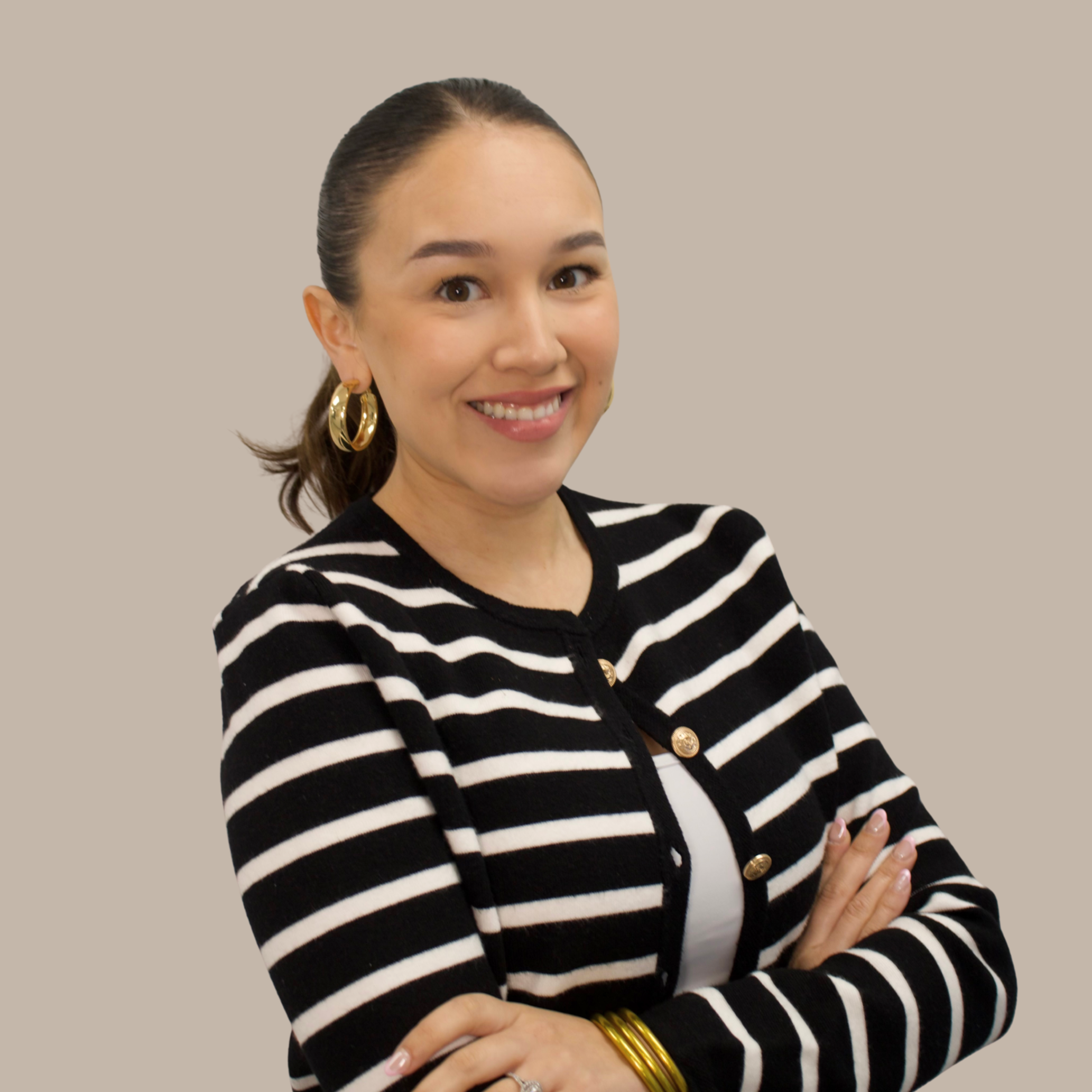 A woman with brown hair tied back, wearing large gold hoop earrings, a black and white striped cardigan with gold buttons, and a white top, smiling with arms crossed against a beige background.