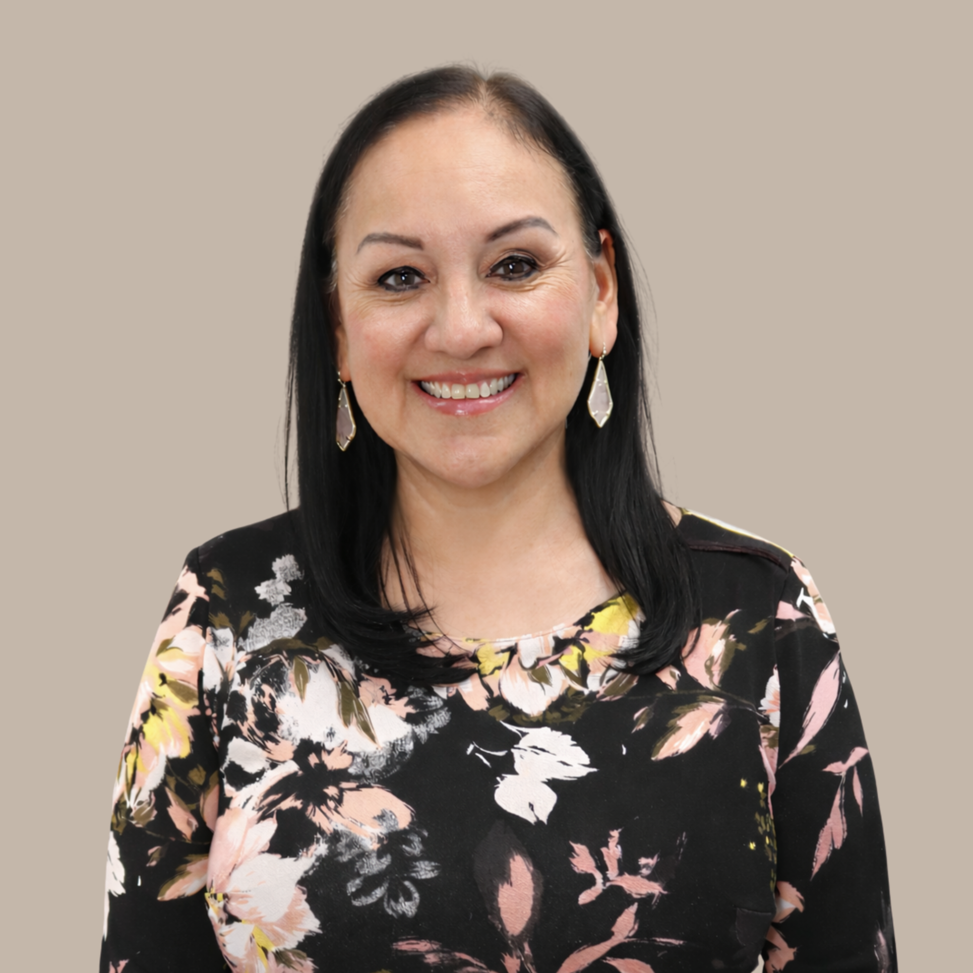 A smiling woman with black hair wearing a floral dress and earrings, standing against a plain beige background.