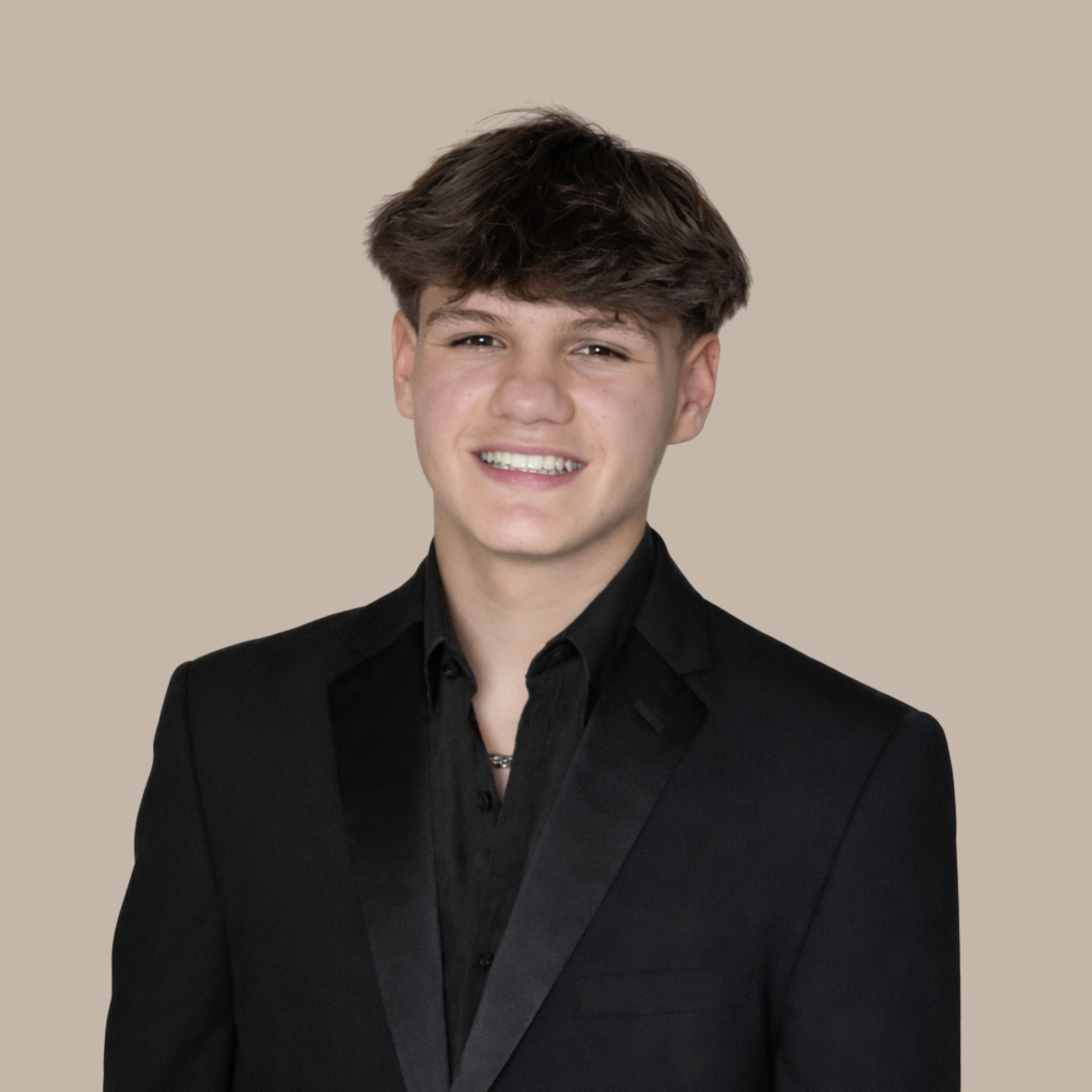 A young man with short brown hair smiling, wearing a black suit with a black shirt, against a plain beige background.