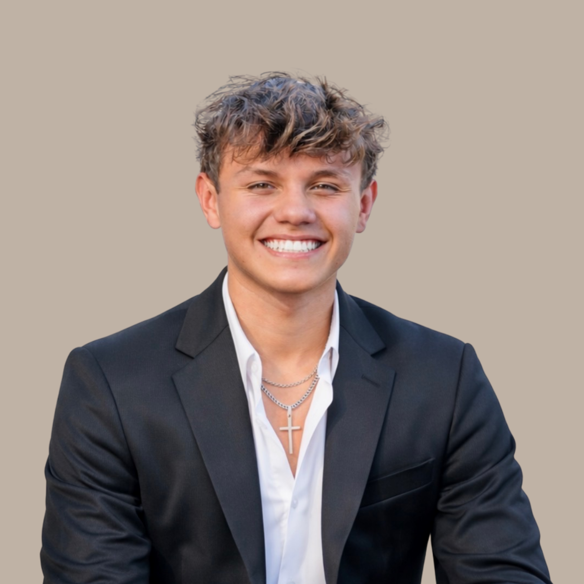 Young man with curly hair wearing a black blazer, white shirt, and silver cross necklace, smiling against a plain beige background.