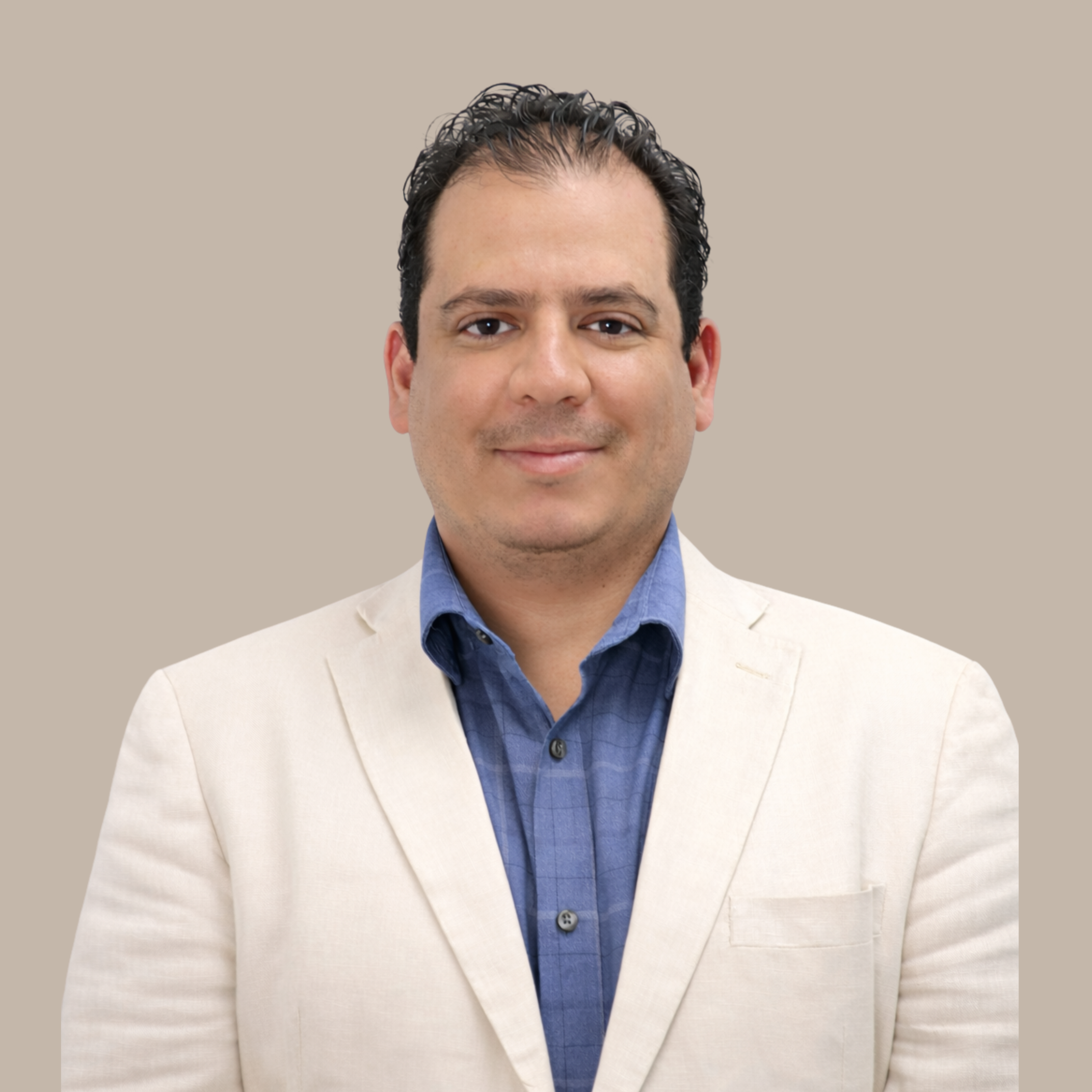 Portrait of a man with dark curly hair, wearing a cream-colored blazer and a blue button-up shirt, smiling softly against a beige background.