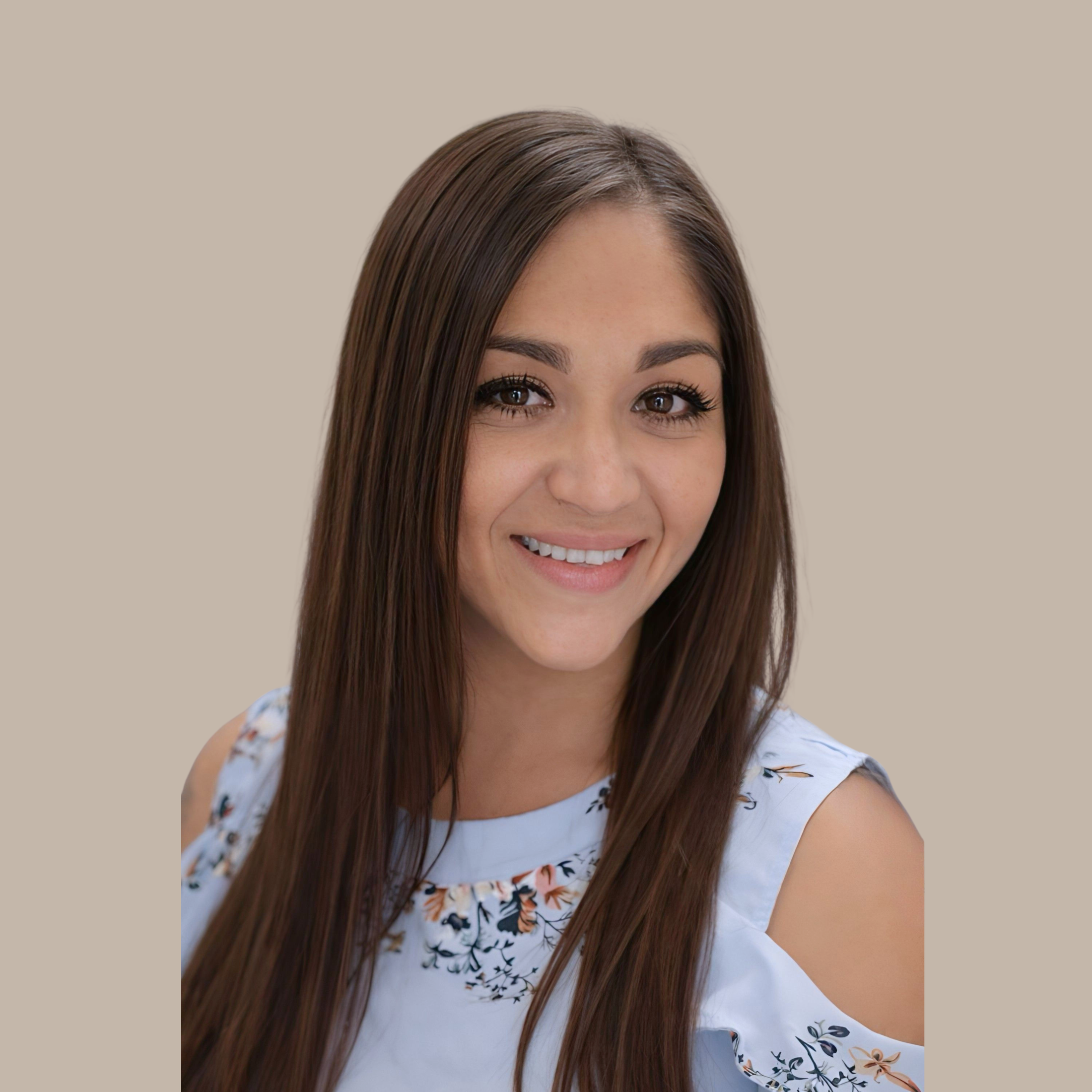 A smiling woman with long brown hair wearing a white floral cold-shoulder top against a beige background.