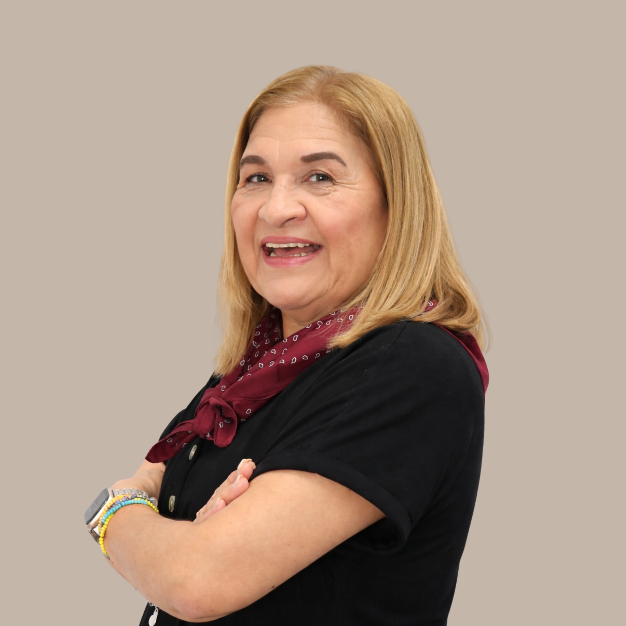 Portrait of a middle-aged woman with blonde hair, smiling, wearing a black shirt and a red bandana, arms crossed, against a neutral background.