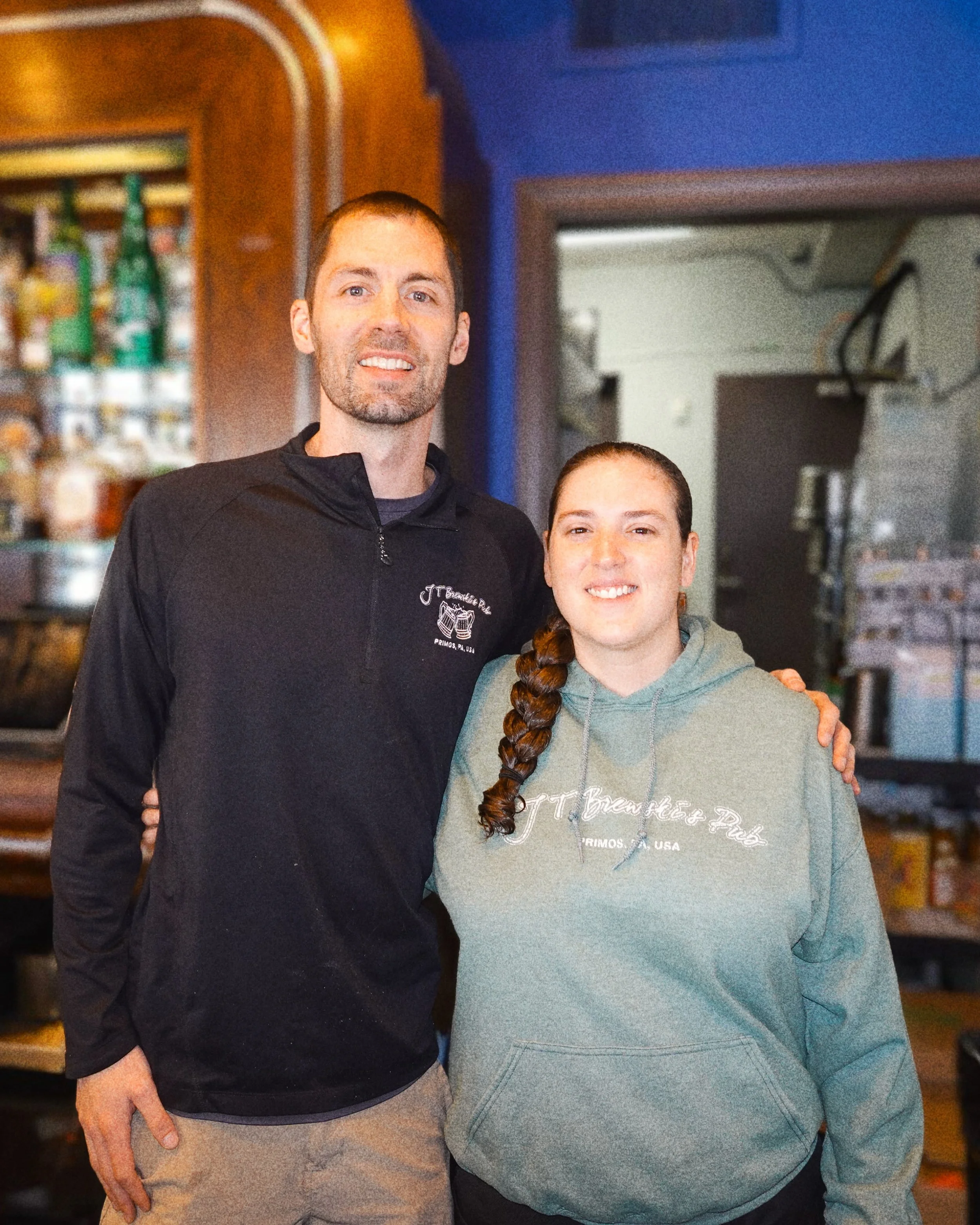 A man and woman standing indoors, smiling, with the man wearing a black hoodie and the woman wearing a gray hoodie, in a room with wooden shelves and a door in the background.