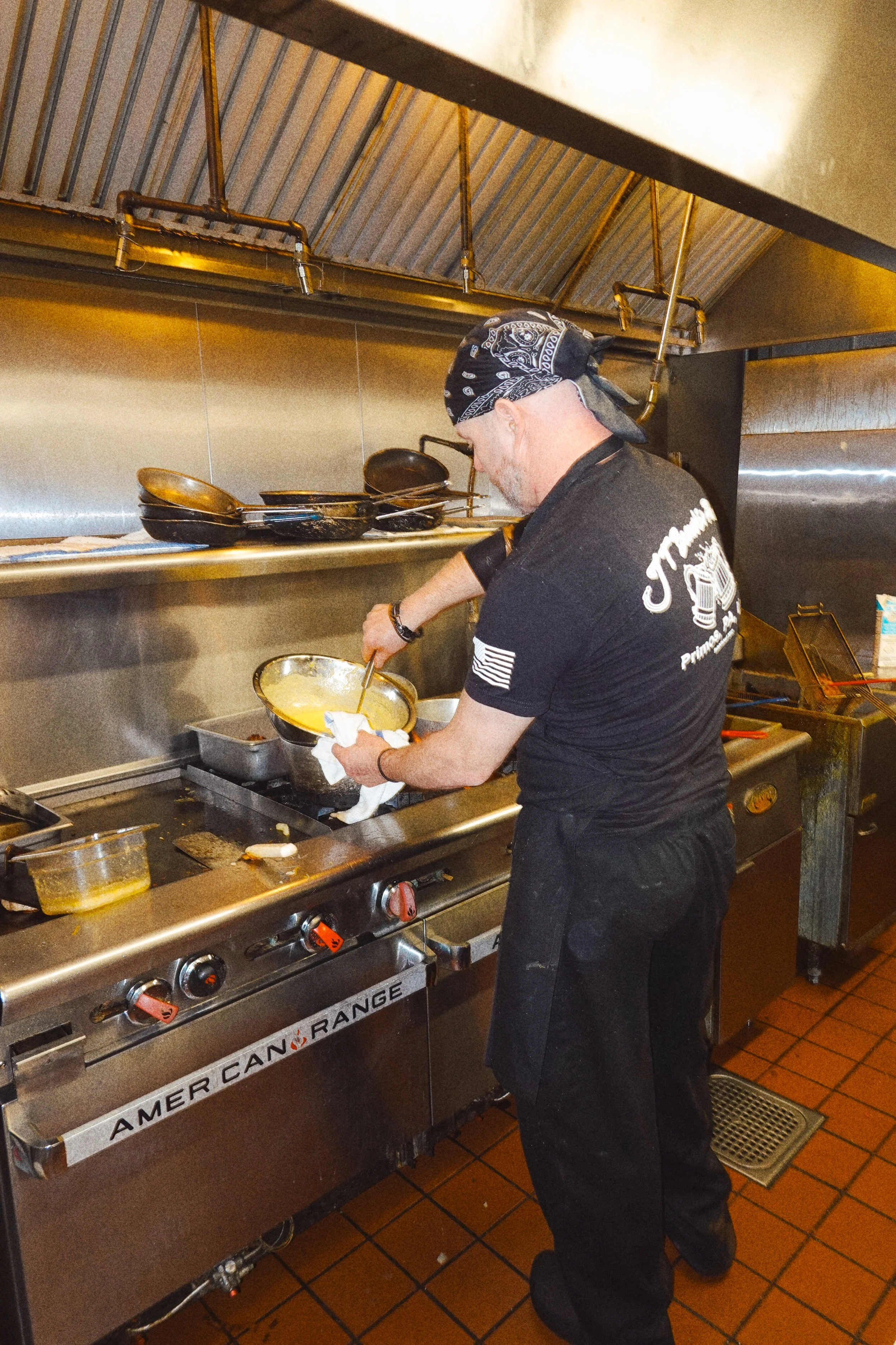 A man wearing a black bandana and shirt is cooking in a commercial kitchen, stirring a bowl over a stove.