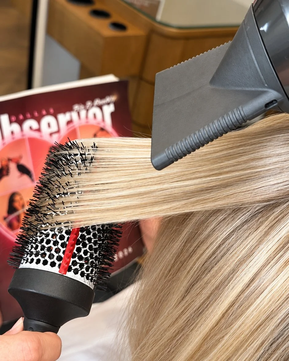 A stylist drying a section of long blonde hair over a round brush with a blow dryer