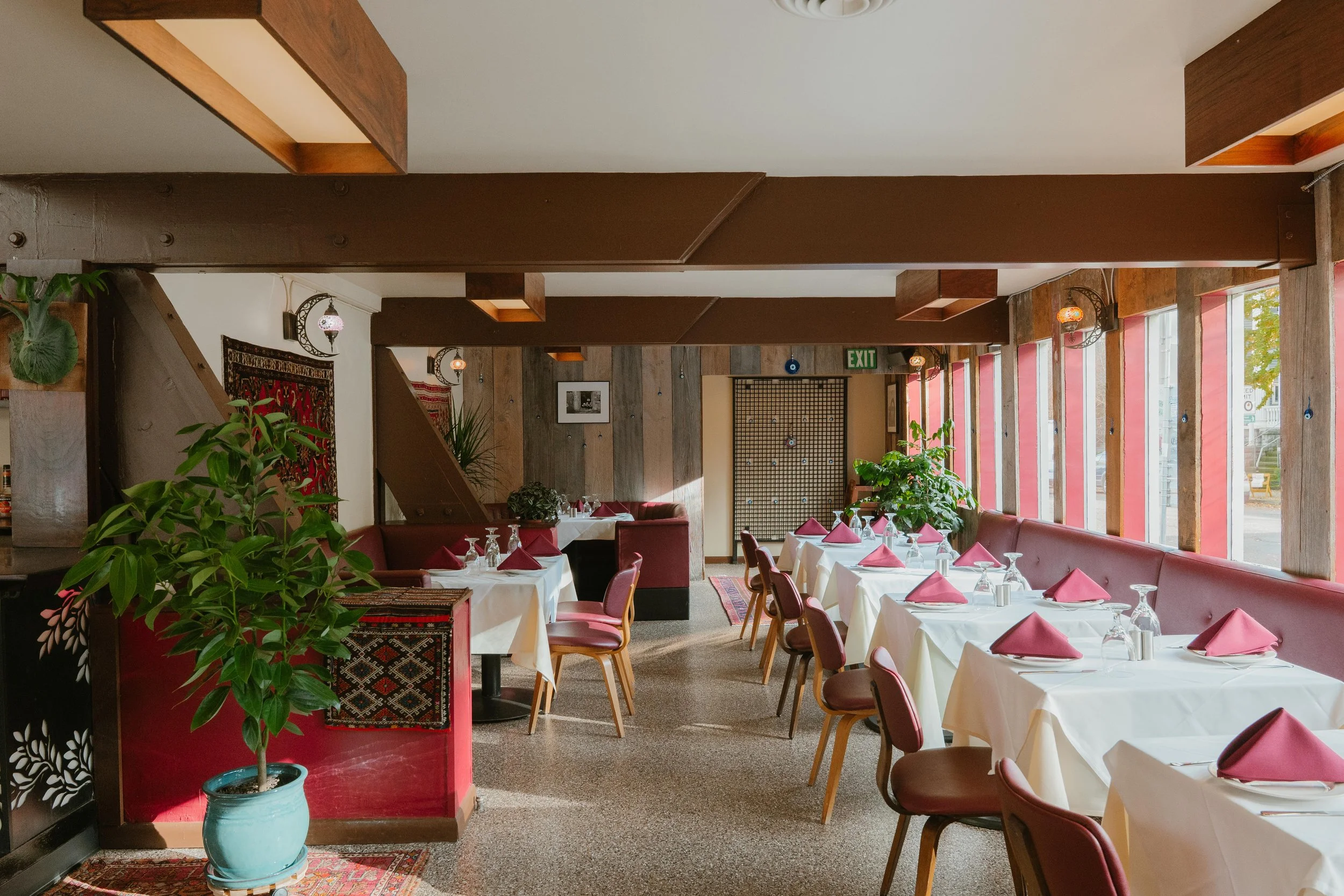 Empty restaurant dining area with white tablecloths, pink napkins, glassware, and pink chairs, decorated with potted plants and wooden accents, illuminated by natural light from large windows.