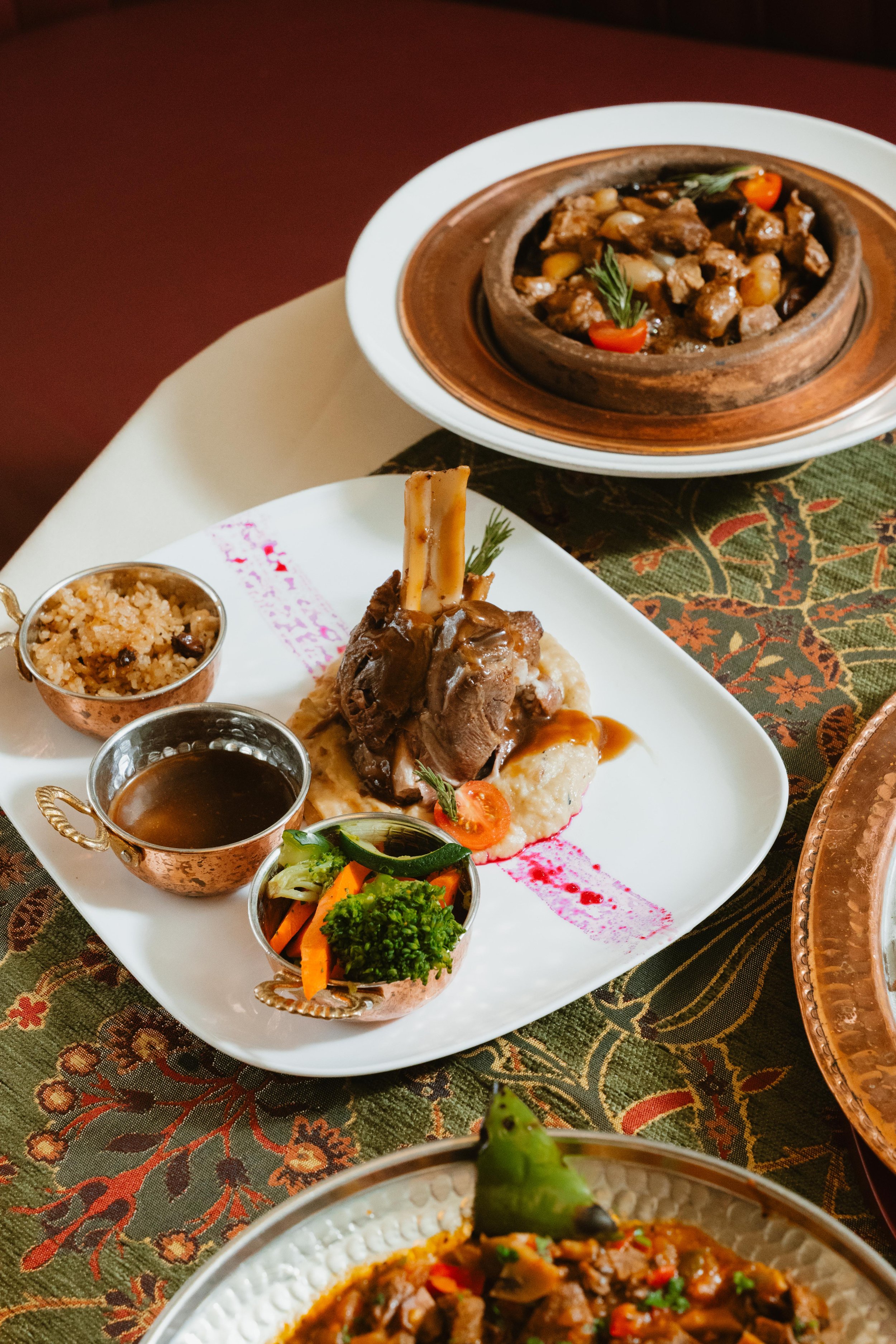 A plate of food with lamb chops, mashed potatoes, and vegetables, alongside small bowls of gravy, vegetables, and rice, on a decorative tablecloth with copper dishes in the background.