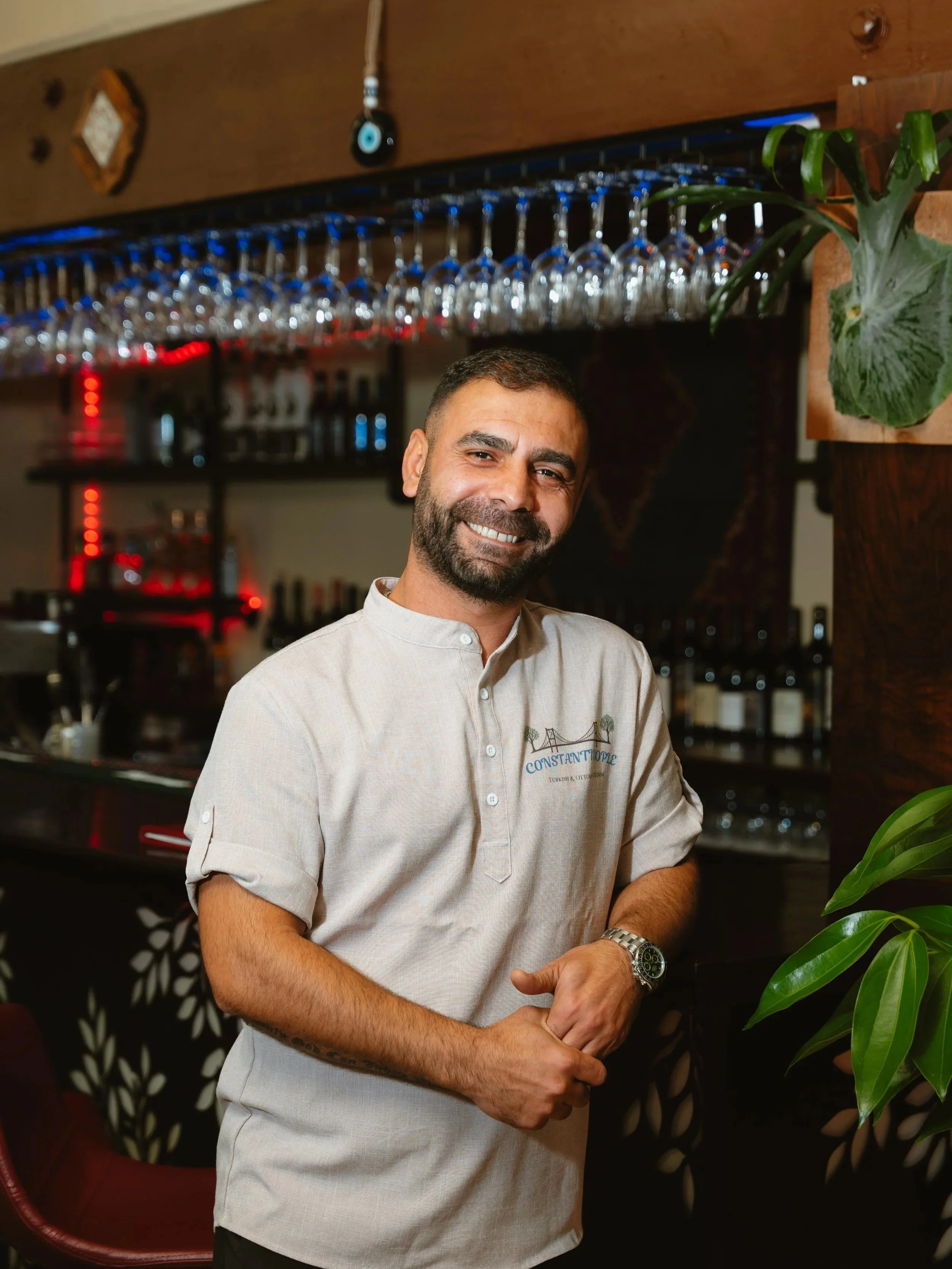 A smiling man with a beard and short hair standing in a bar or restaurant, wearing a beige shirt with a logo, among plants and glassware.