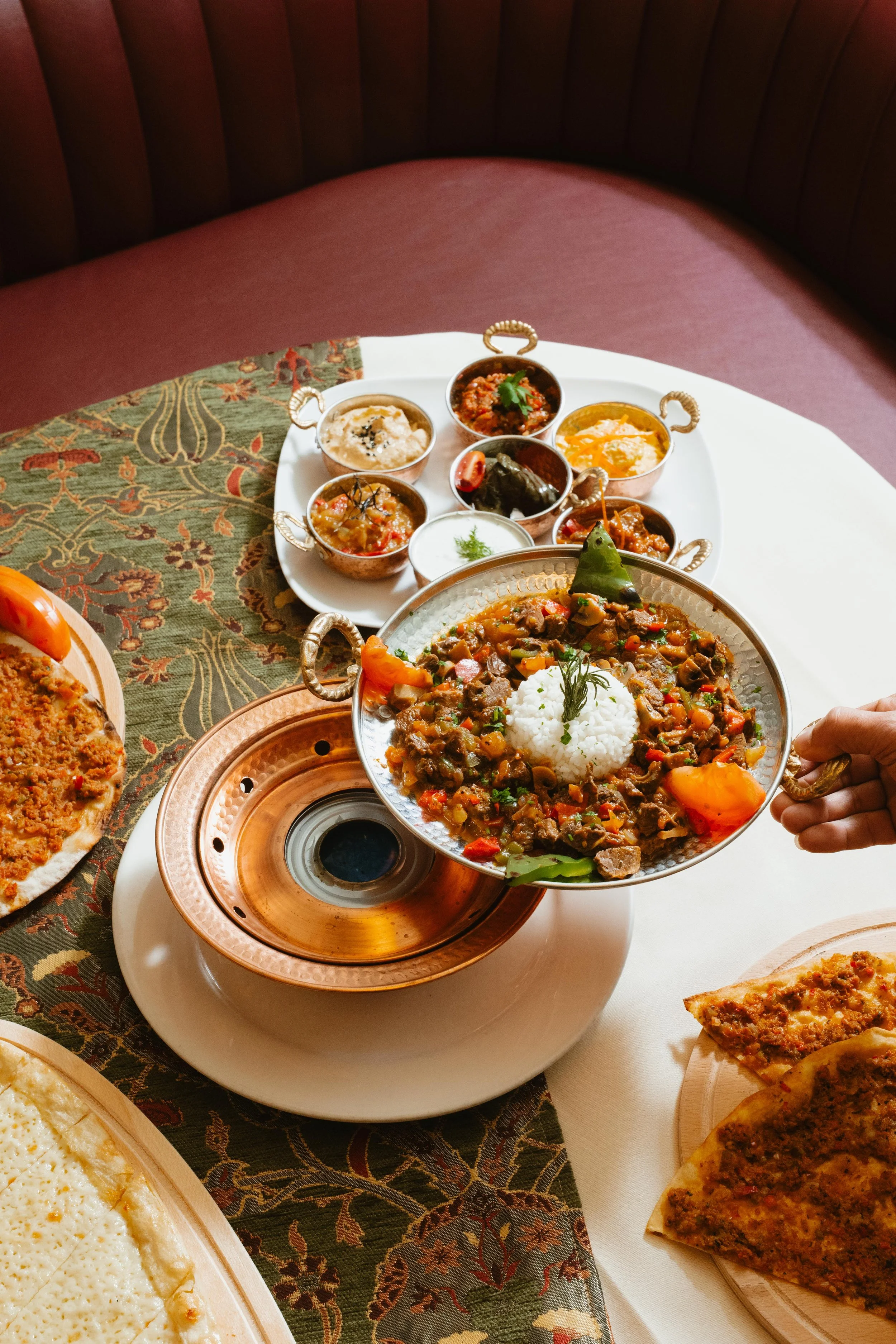 A hand holding a silver serving tray with rice and vegetable curry, surrounded by various Indian dishes and breads on a white table.