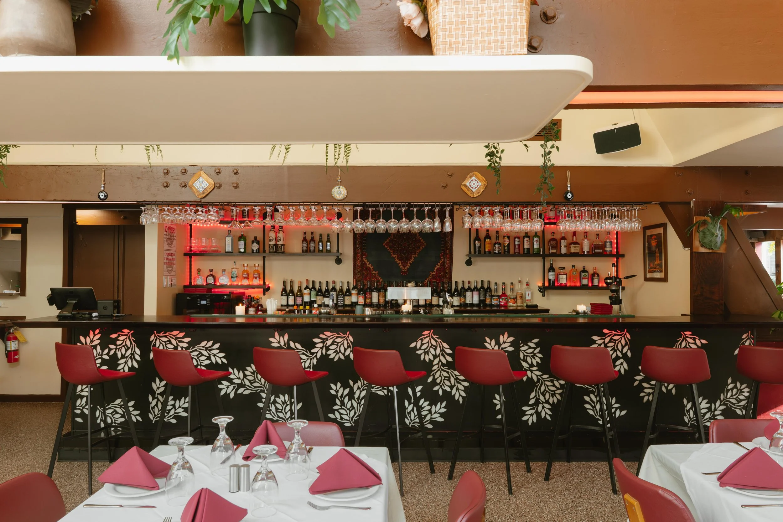 Indoor restaurant bar with red chairs, a black bar counter with a white leaf pattern, and a well-stocked bar area with bottles and glassware, decorated with plants and framed pictures.