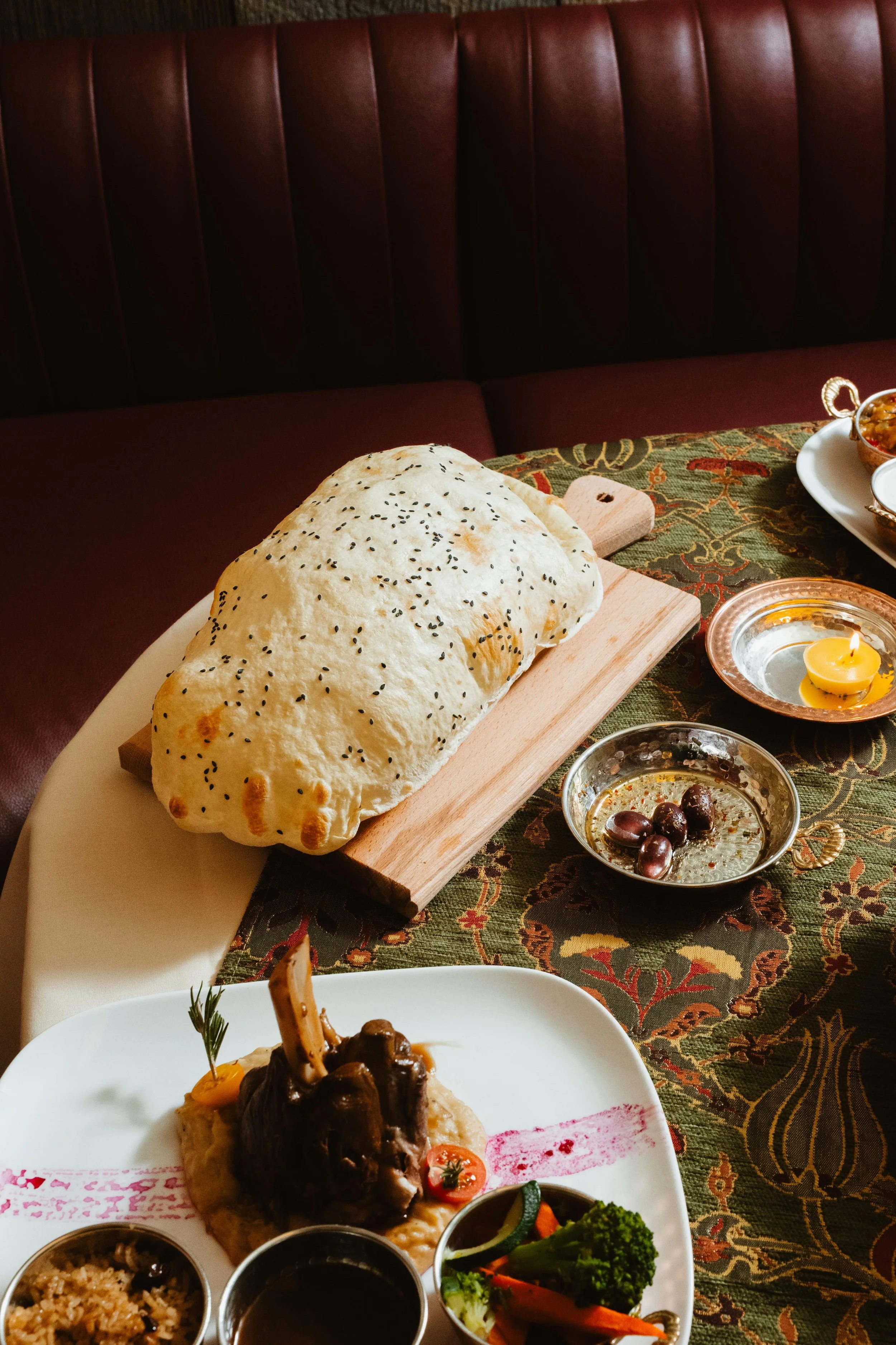 A loaf of bread with black sesame seeds on a wooden cutting board, restaurant setting with a patterned tablecloth, dishes with curry, vegetables, and various sauces.