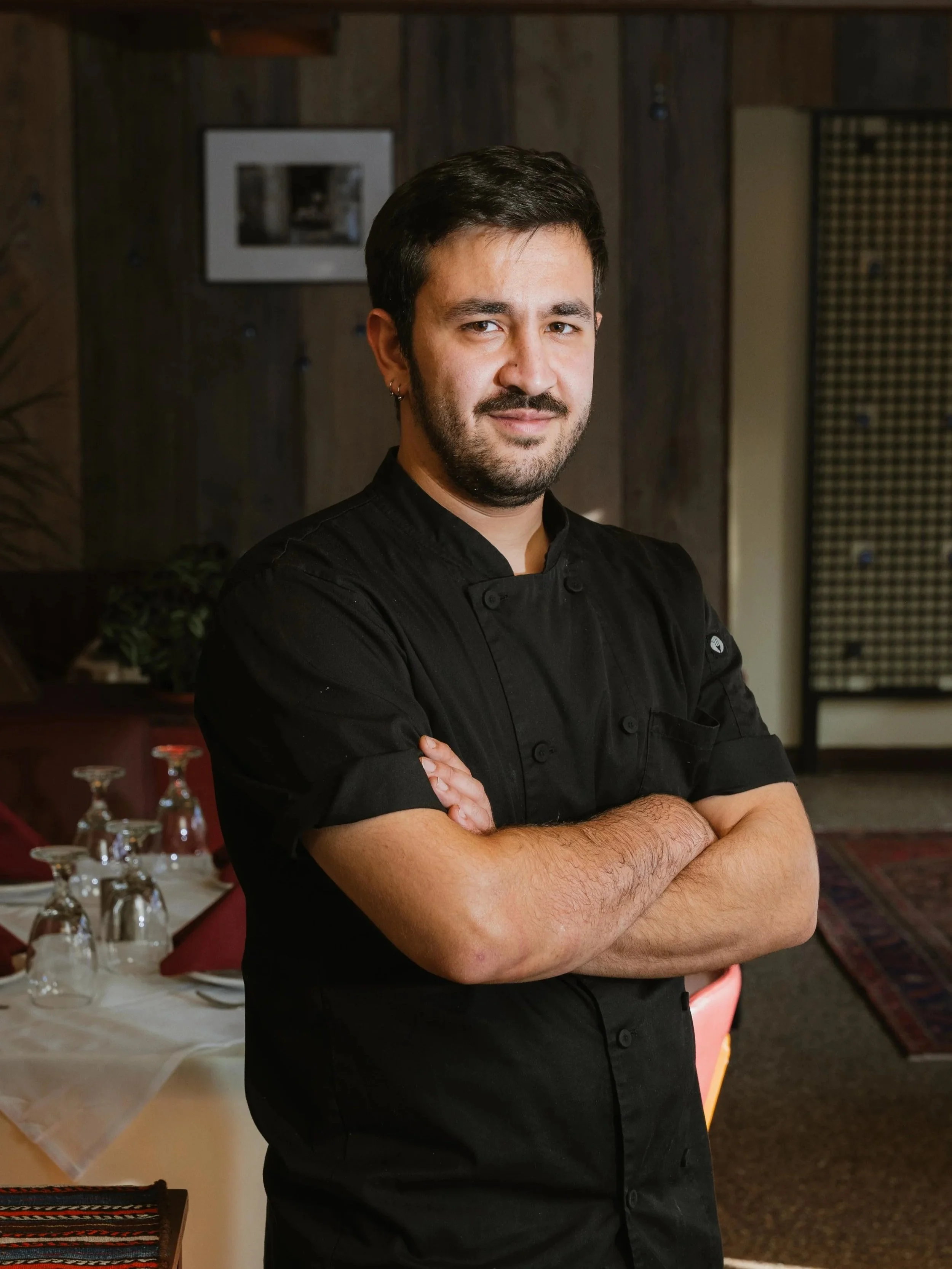 A man in a black chef's coat standing with arms crossed, in a restaurant setting with tables, glasses, and a wooden wall background.