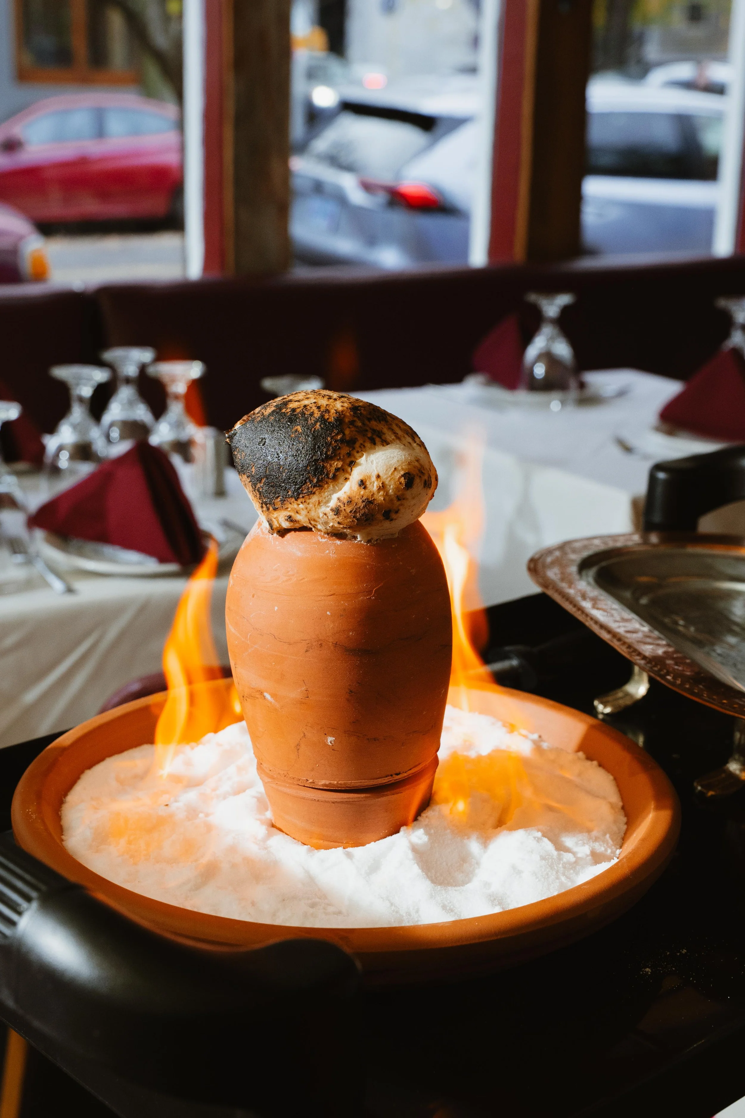 A Molten Chocolate Margarita being prepared at a restaurant, with flames surrounding it on a table with white tablecloths and wine glasses, in a restaurant setting with windows showing parked cars outside.