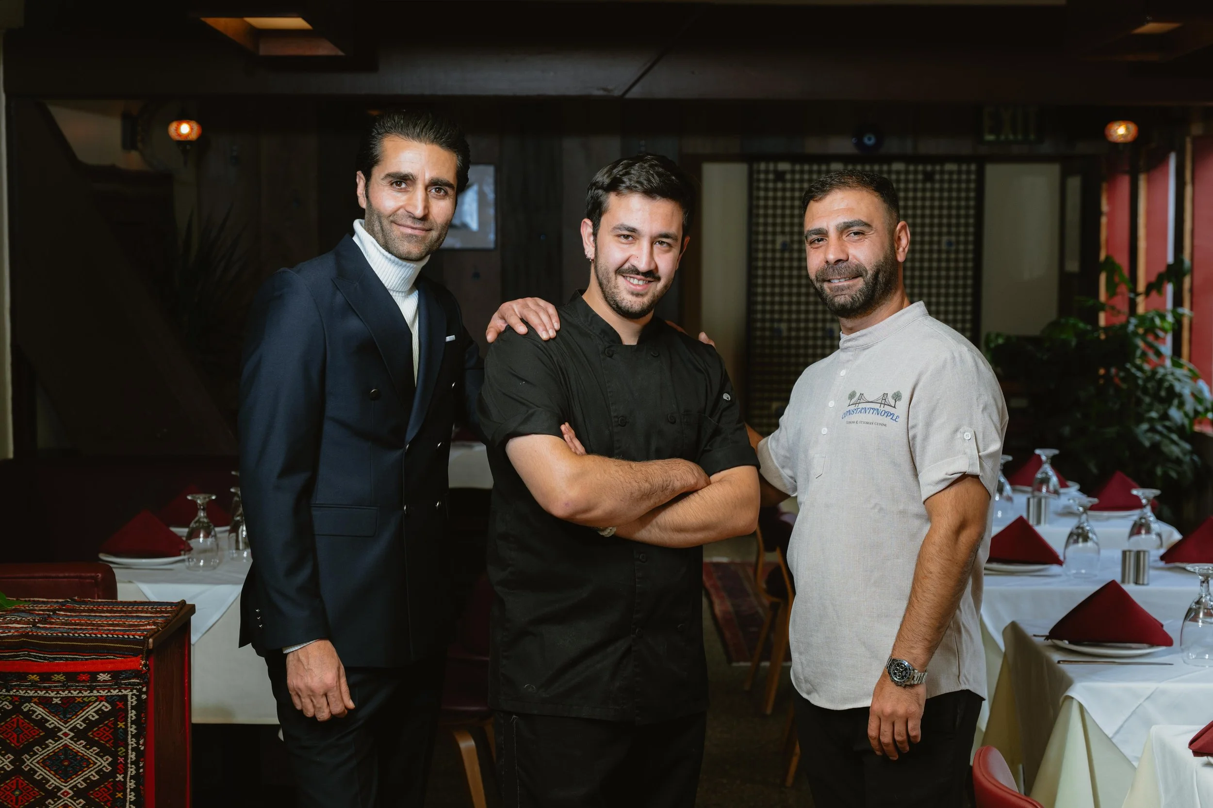 Three men standing together in a restaurant with tables set for dining, smiling at the camera.