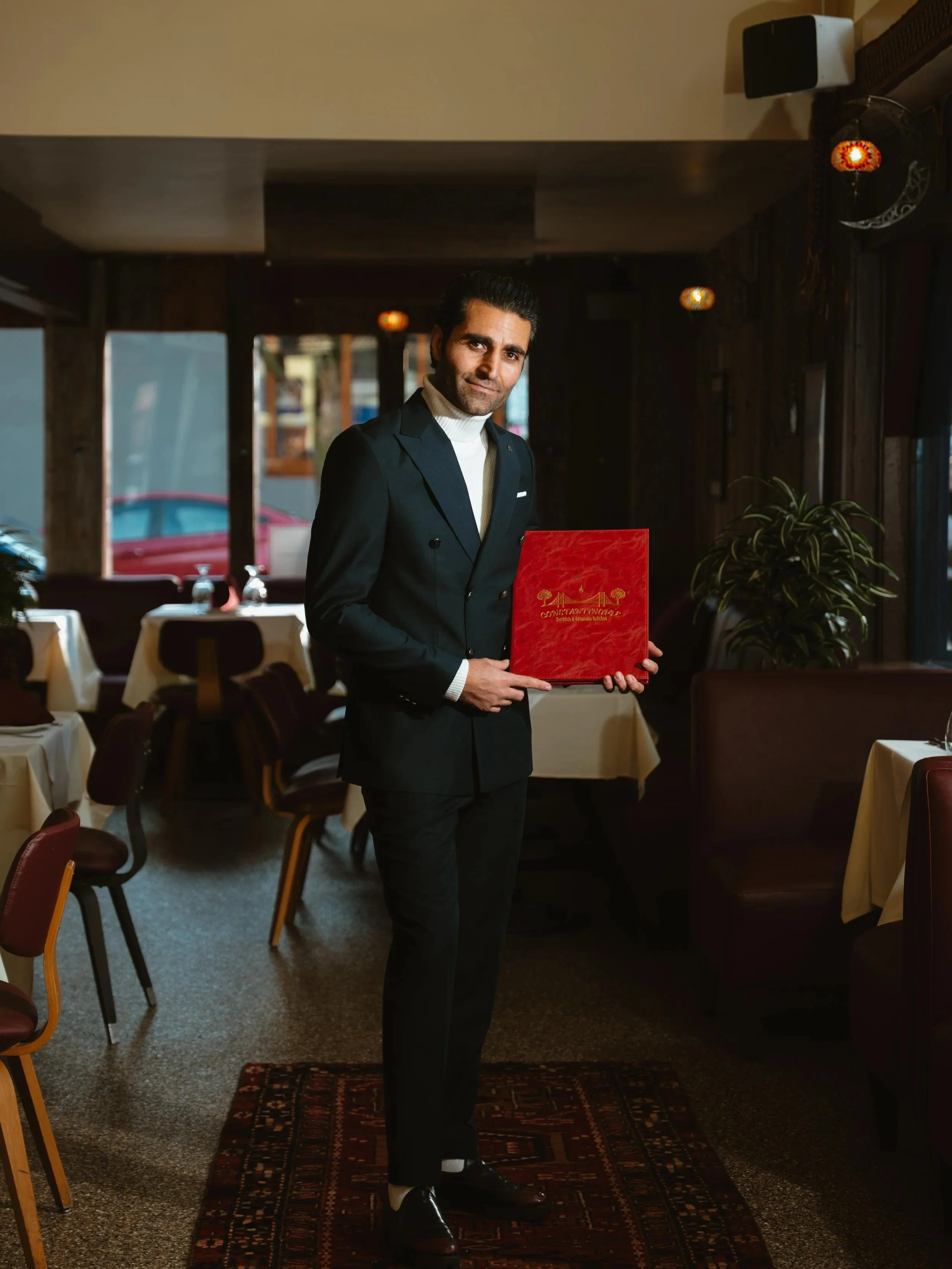 A man in a black suit holding a red menu in a restaurant with dining tables and chairs, dim lighting, and decorative wall lamps.