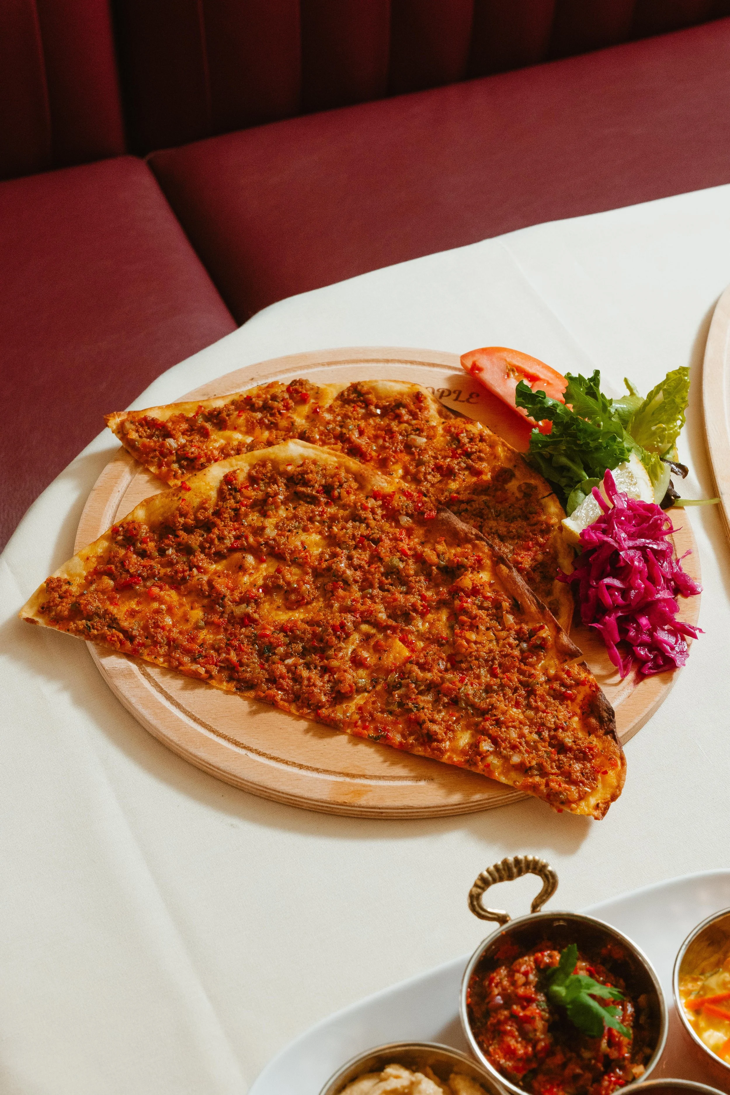Sliced pizza with ground meat topping on a wooden serving board, garnished with a side of lettuce, tomato, and purple cabbage, placed on a white tablecloth.
