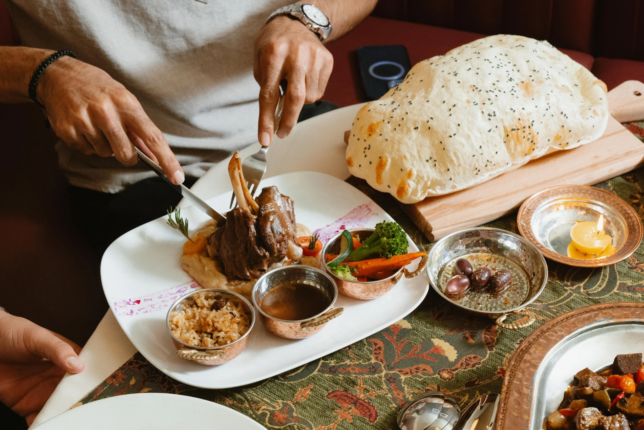 Person serving a lamb shank with mashed potatoes and vegetables at a festive dinner table. There is a large loaf of bread on a wooden board, candles, and various dishes with sauce, rice, and salad in copper bowls on the table with a patterned tablecloth.