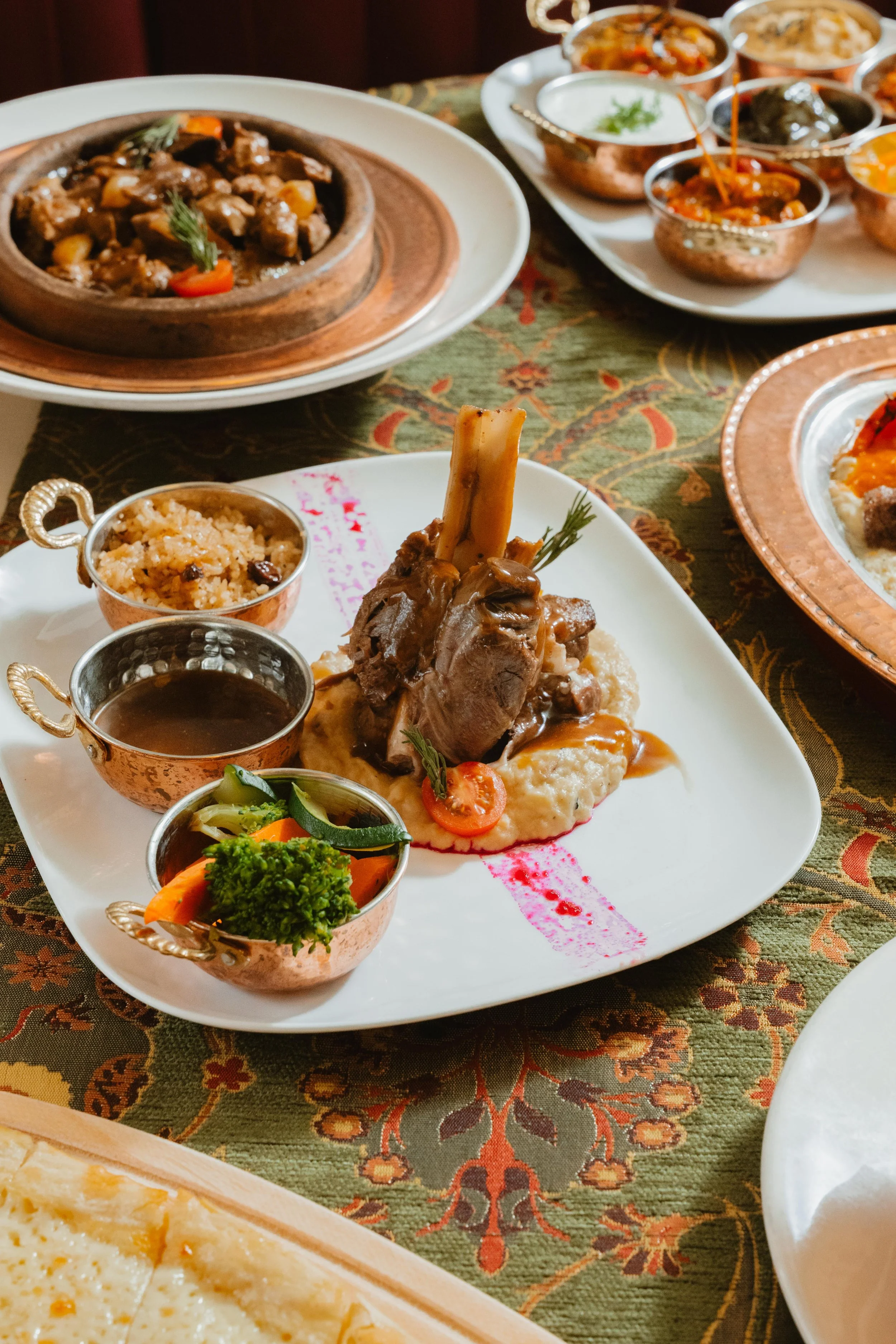 A plated meal featuring a large lamb chop served with mashed potatoes, garnished with cherry tomato slices, accompanied by three small bowls of sauce and vegetable medley, on an ornate tablecloth with other dishes in the background.