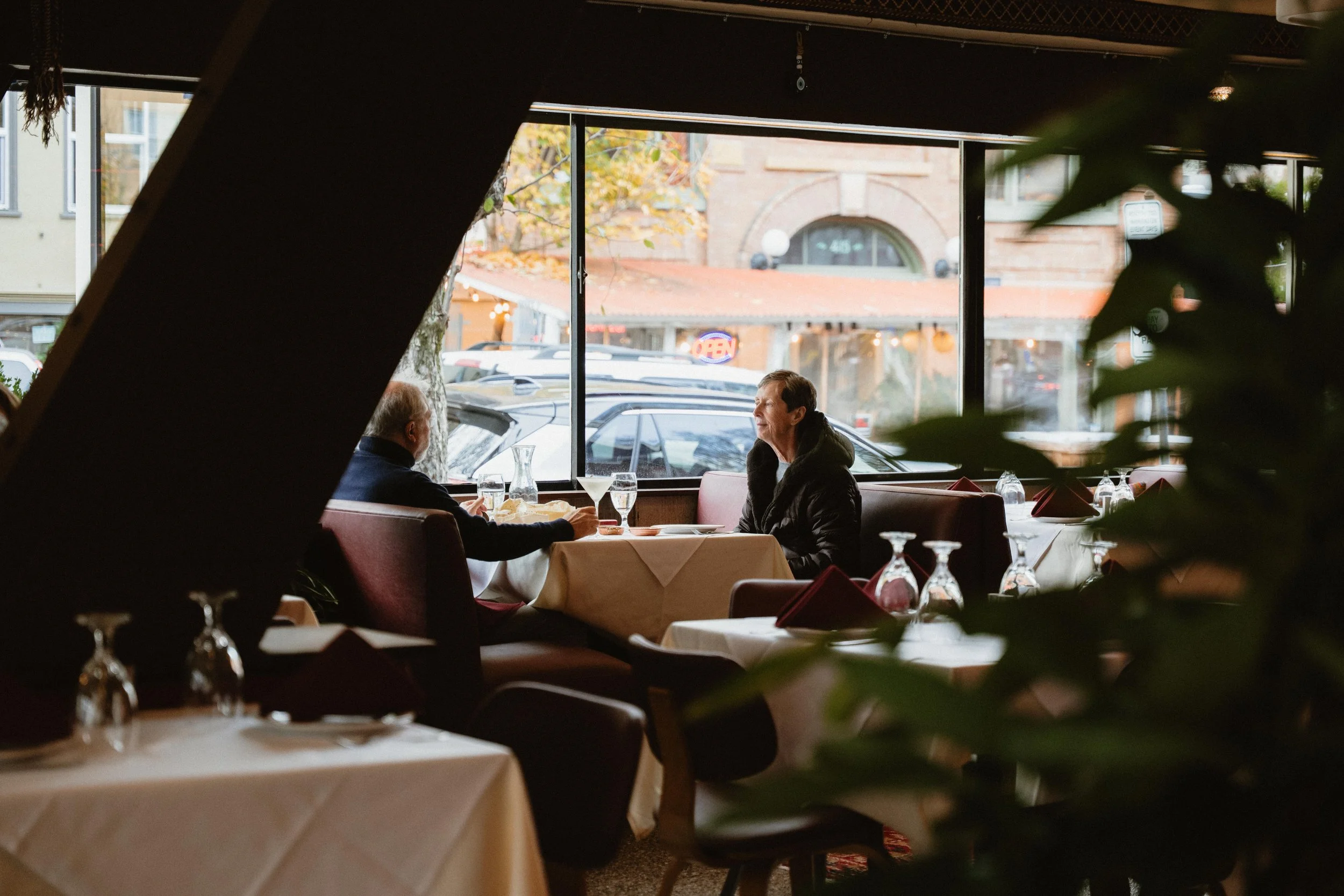 Two men are sitting at a table in a restaurant with large window views outside, with parked cars and a building in the background. The table is set with glassware and napkins, and the restaurant's interior features white tablecloths and dim lighting.