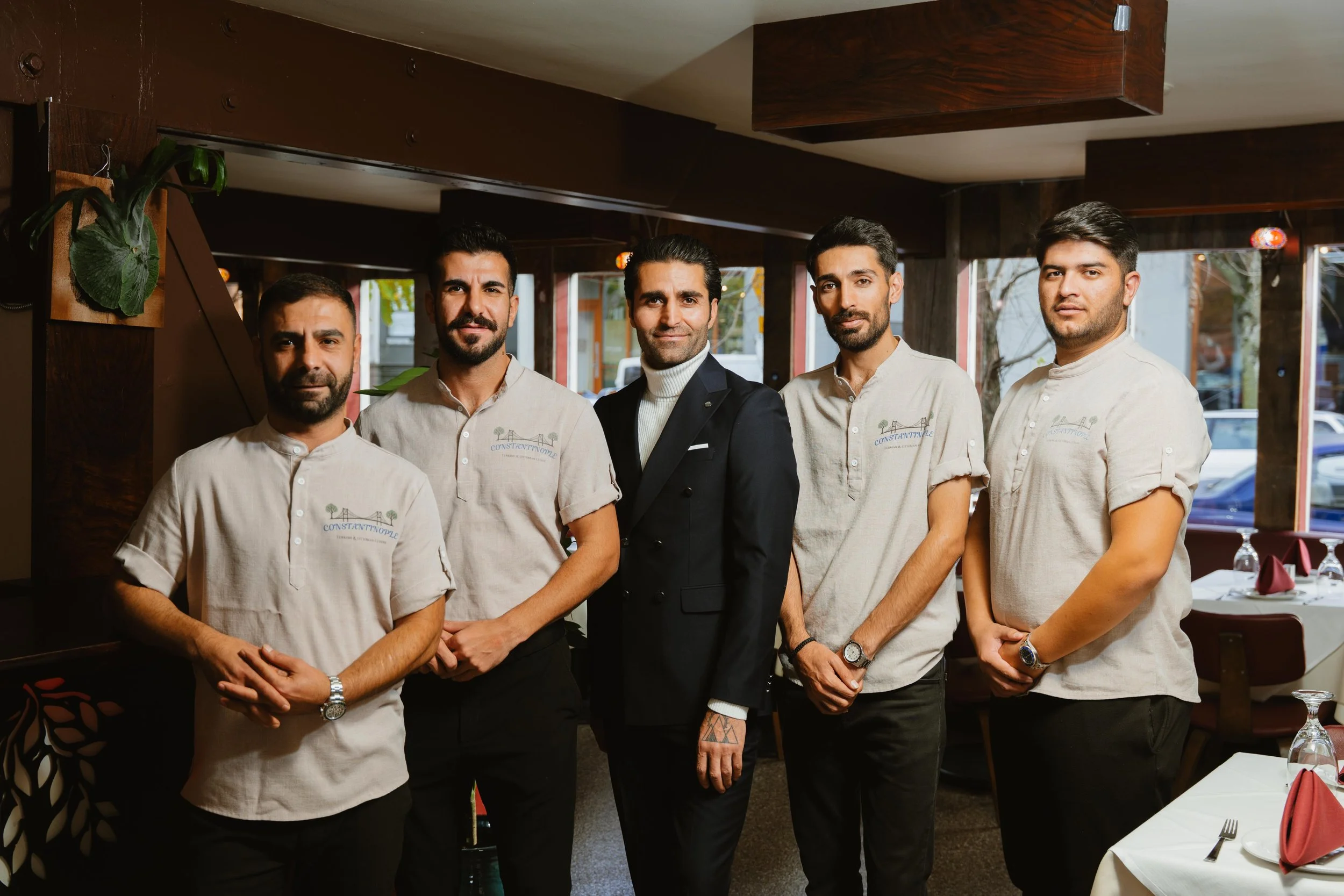 Group of five men in a restaurant, four wearing beige shirts with embroidered logos, one in a black suit, standing indoors near windows, dining tables set with napkins and glasses.