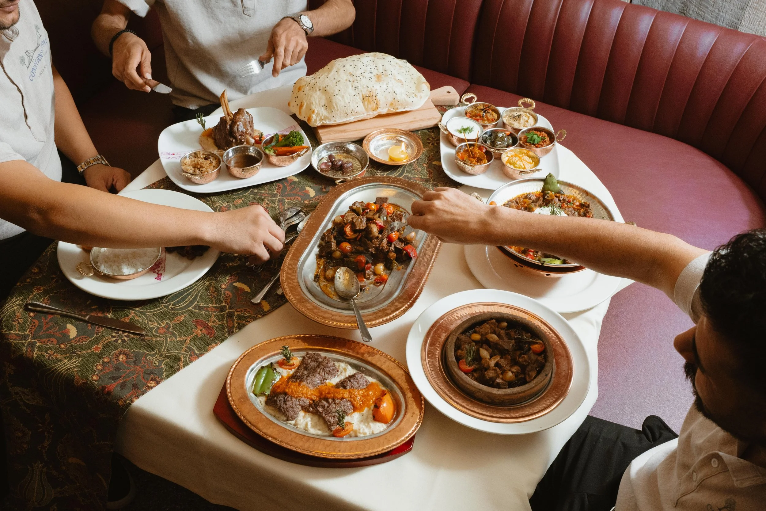A group of people enjoying a meal with various Middle Eastern dishes on a table, including a large flatbread, stews, and small bowls of sauces and condiments.