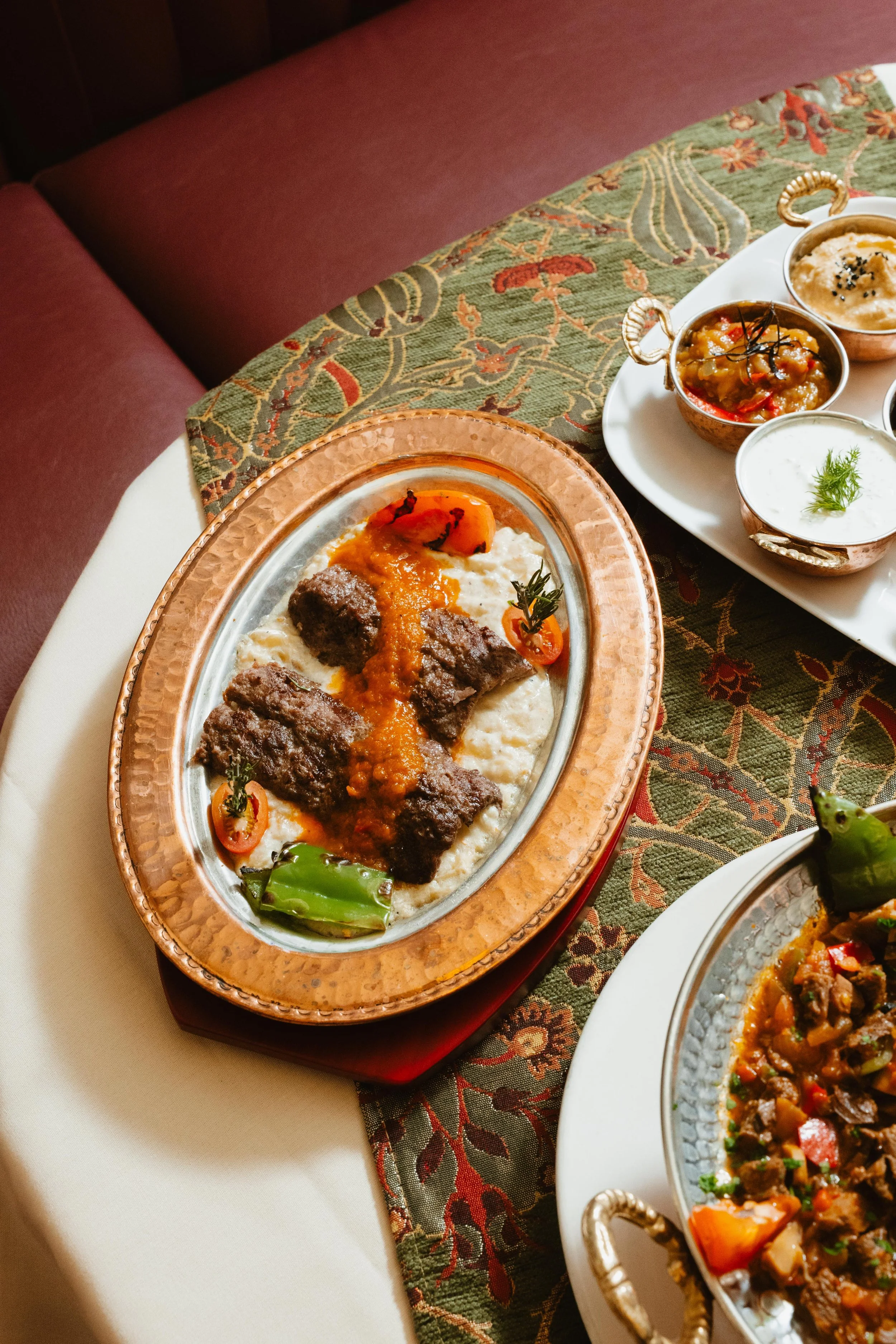 A serving platter with grilled meat, mashed potatoes, cherry tomatoes, and green peppers on a table with a patterned tablecloth and other dishes of food.