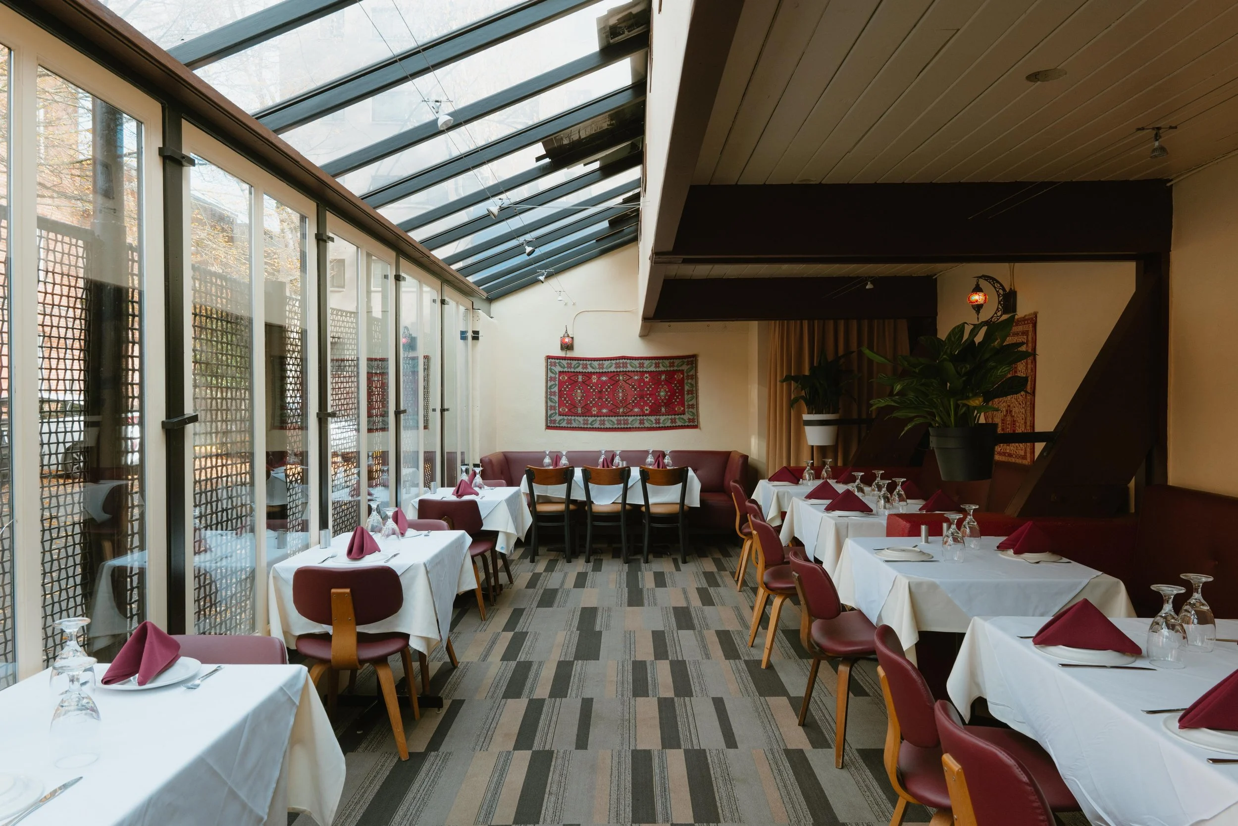 Interior of a restaurant dining area with tables set with white tablecloths, maroon cloth napkins, upside-down wine glasses, and utensils. There are large glass windows on the left letting in natural light, and a decorative tapestry on the wall. Potted plants are placed on a partition and a shelving unit, with warm beige walls and a wooden ceiling.