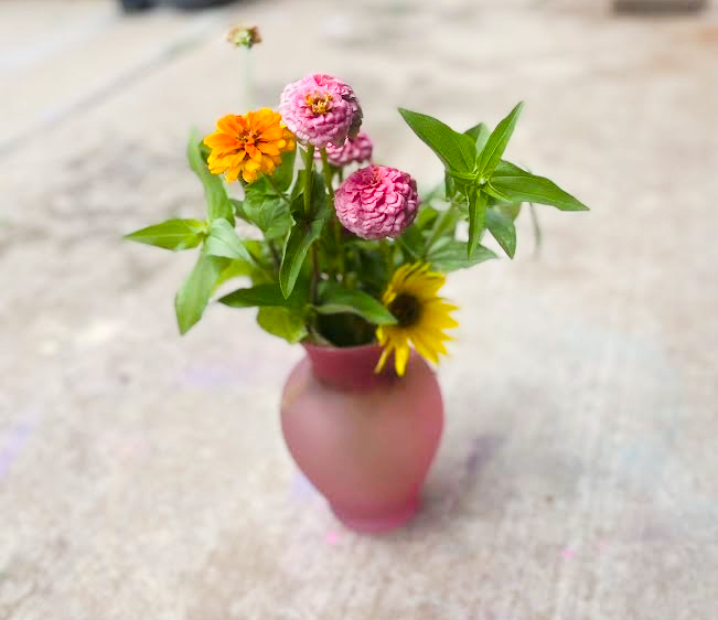 A pink vase holding a small bouquet of colorful flowers, including pink, yellow, and orange blooms with green leaves, on a neutral surface.