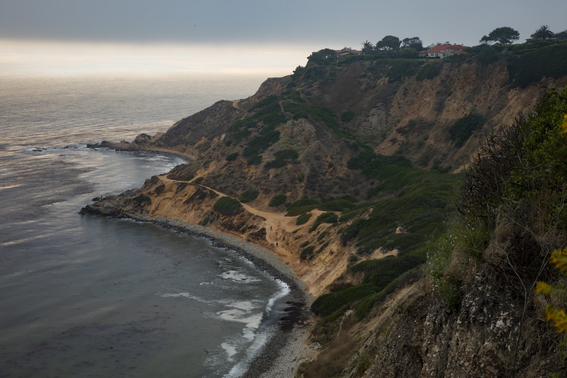 Cliffside coastal landscape with a dirt path, houses, and trees at the top of the cliffs, overlooking the ocean on a partly cloudy day.