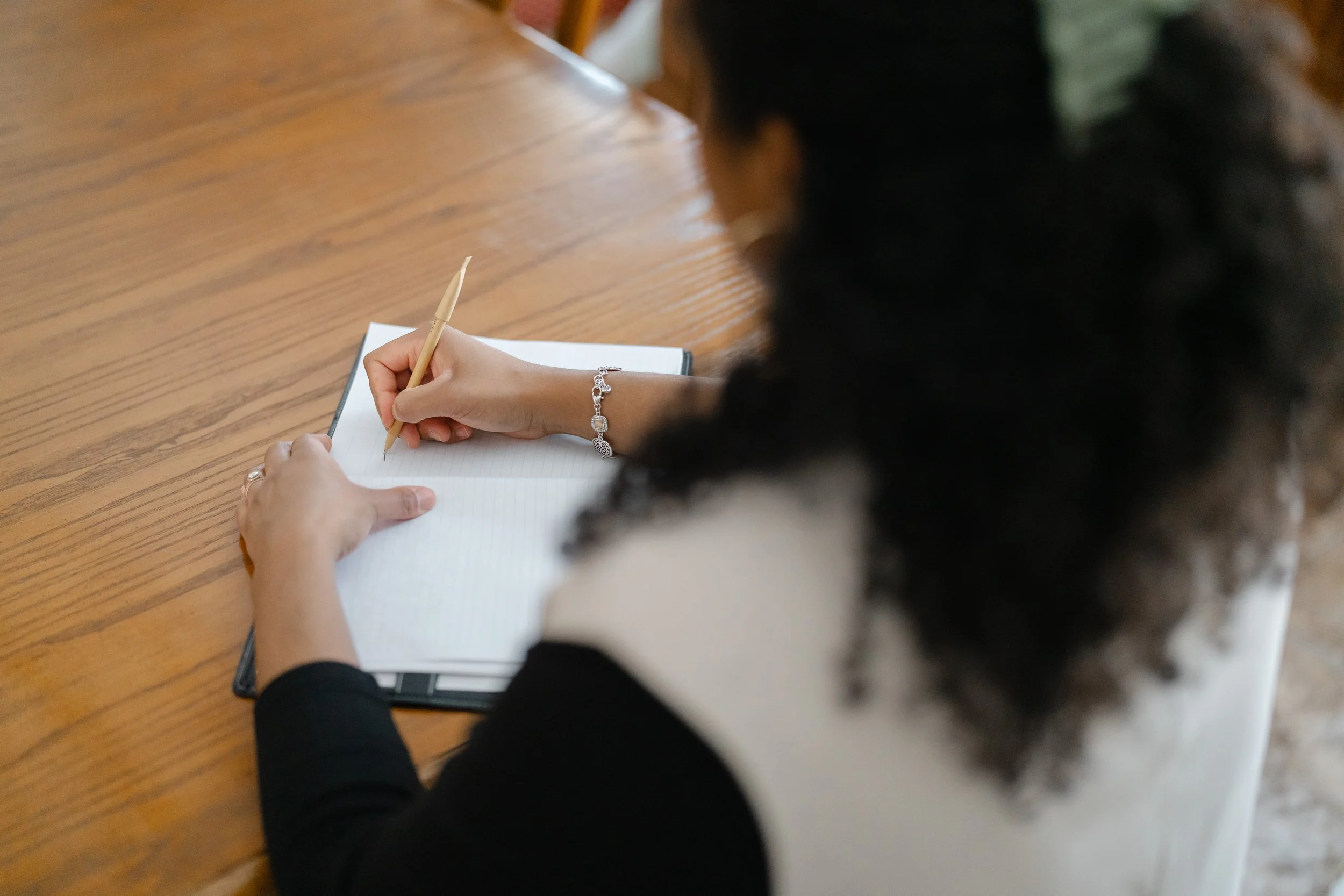 A person with dark, curly hair wearing a bracelet, writing in a notebook with a wooden pen on a wooden table.