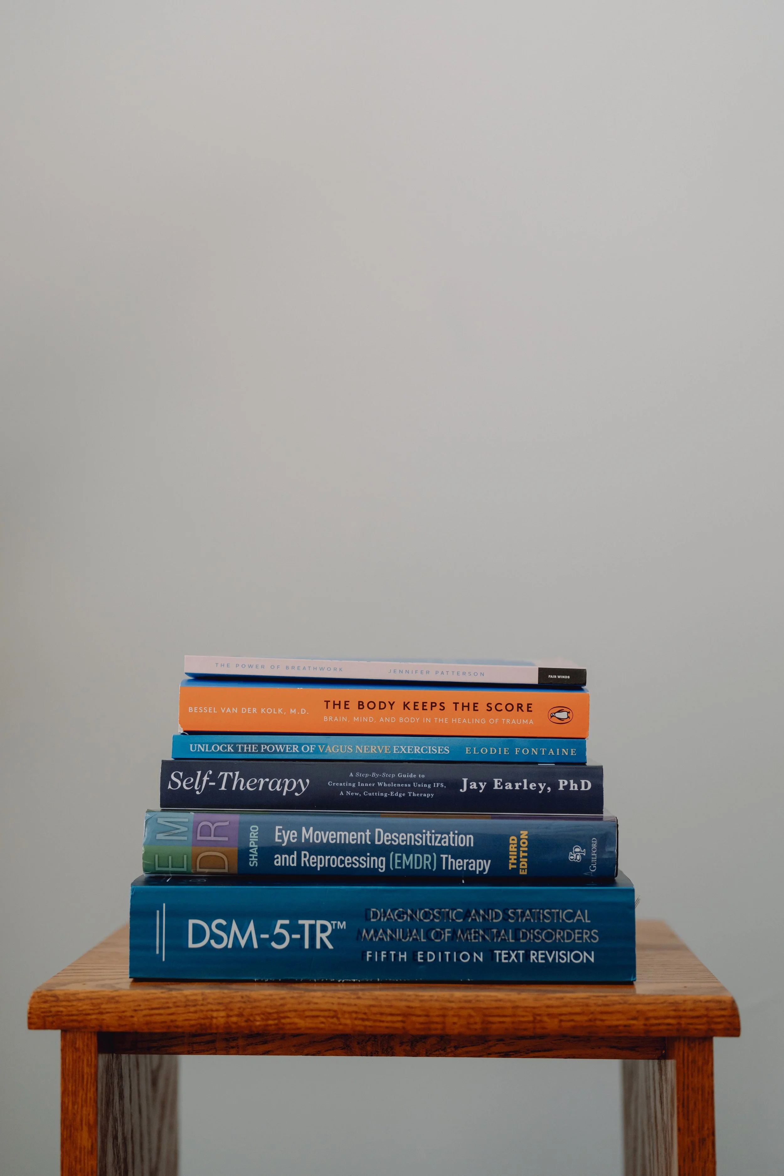Stack of eight psychology and therapy books on a wooden table against a gray wall.