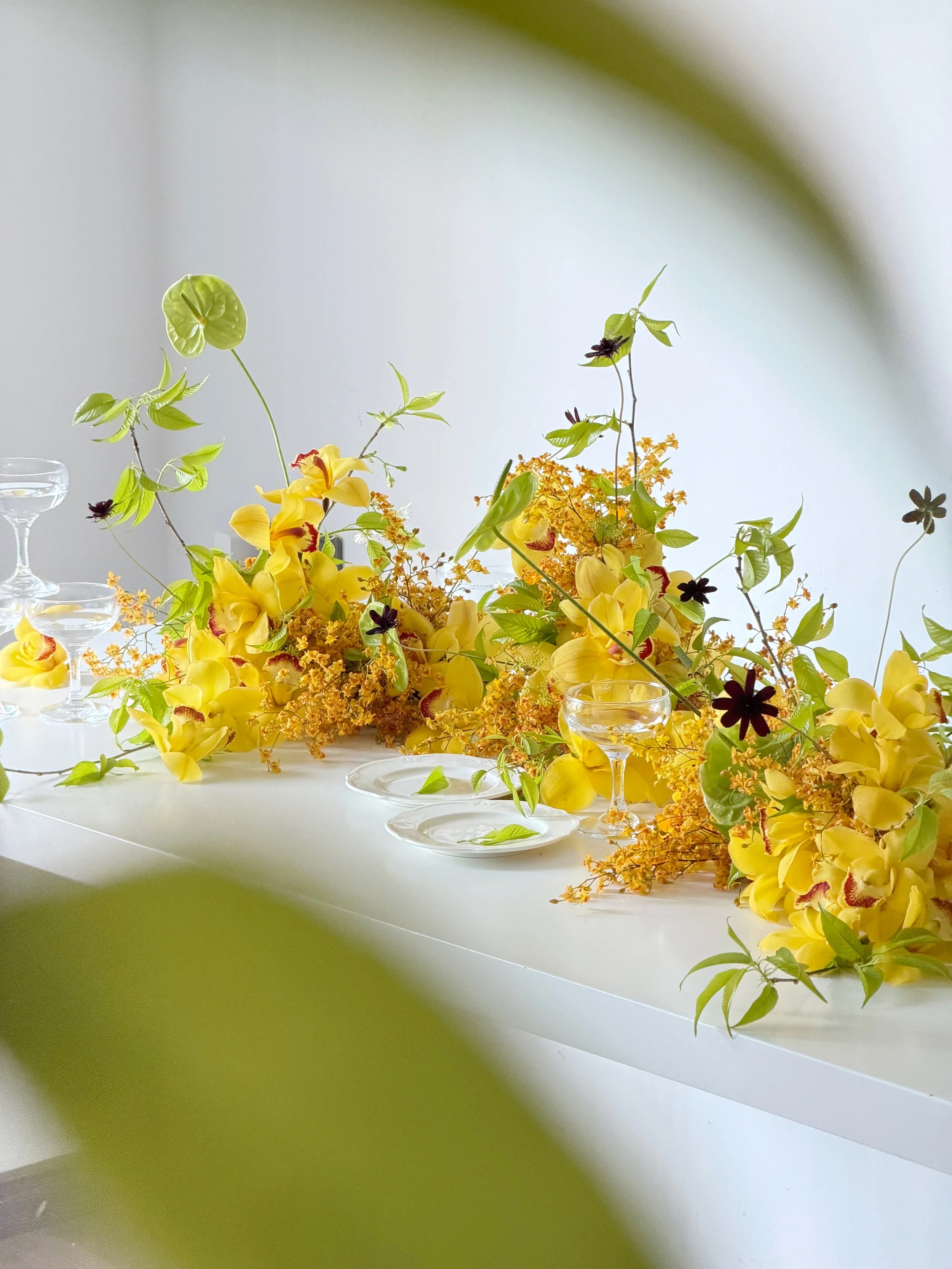 A floral arrangement featuring yellow and dark purple flowers, and green leaves on a white table with glassware, with a plain light-colored wall background.