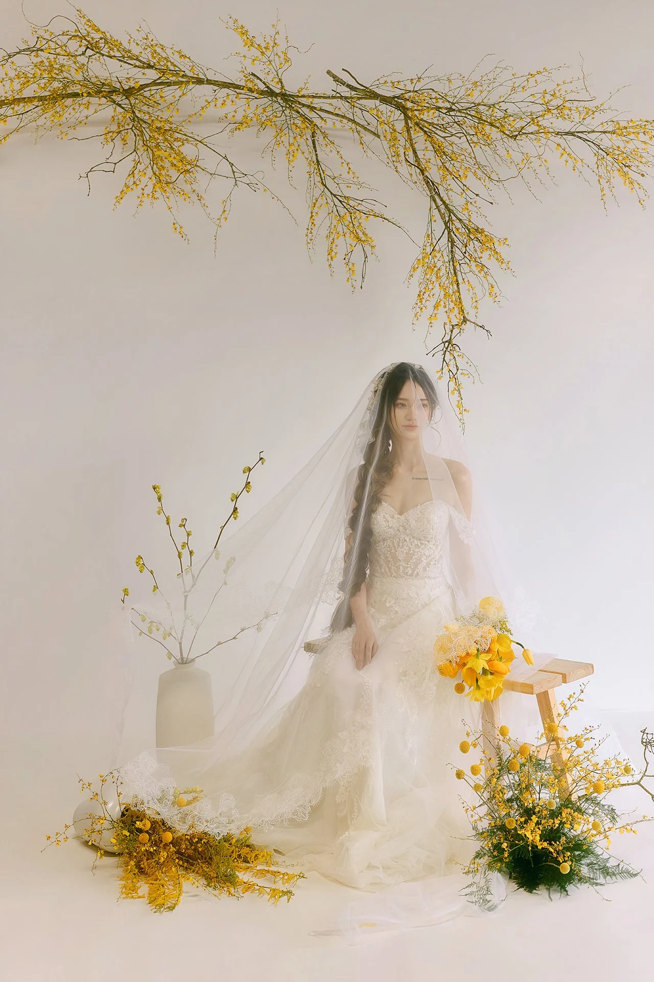 A woman in a wedding dress with lace details, sitting against a plain background, wearing a veil. She is surrounded by yellow flowers and branches, with a vase of yellow blossoms on her left and a wooden stool with more yellow flowers on her right.