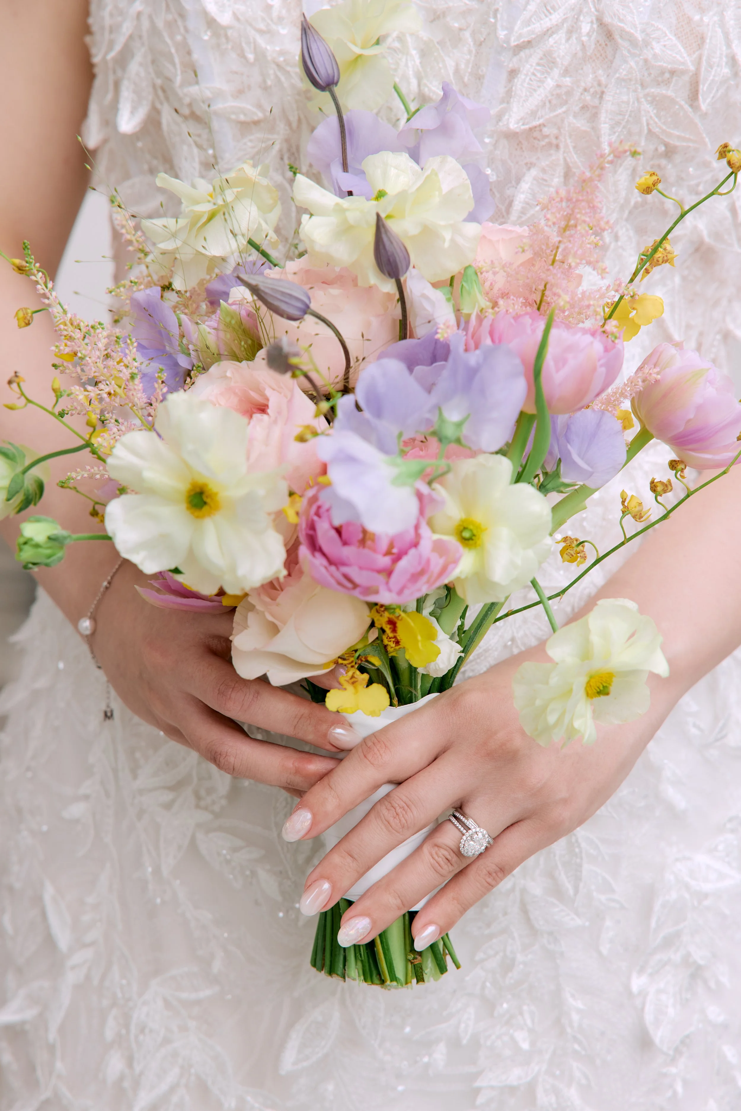 Close-up of a bride holding a bouquet of pastel-colored flowers, including pink, purple, white, and yellow blooms, with her hands and wedding ring visible, against a lace wedding dress background.
