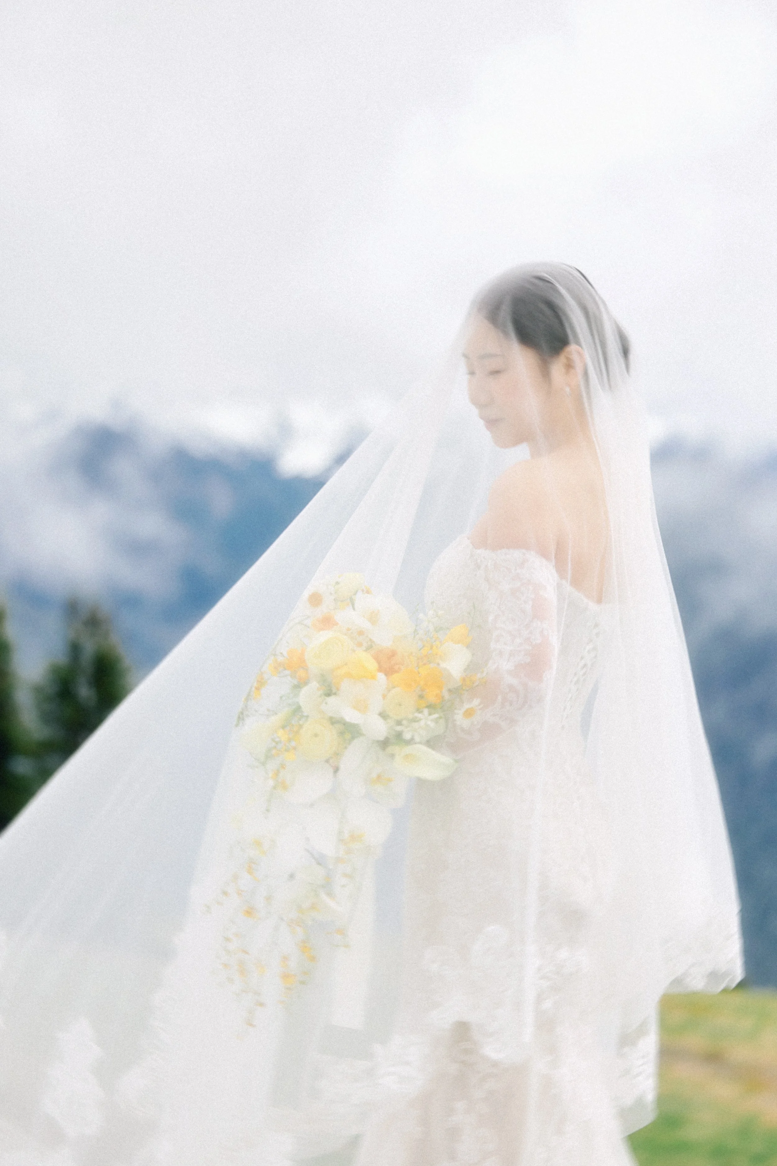 A bride in a white wedding dress holding a bouquet of yellow and white flowers, with a veil over her face, standing outdoors with mountains in the background.
