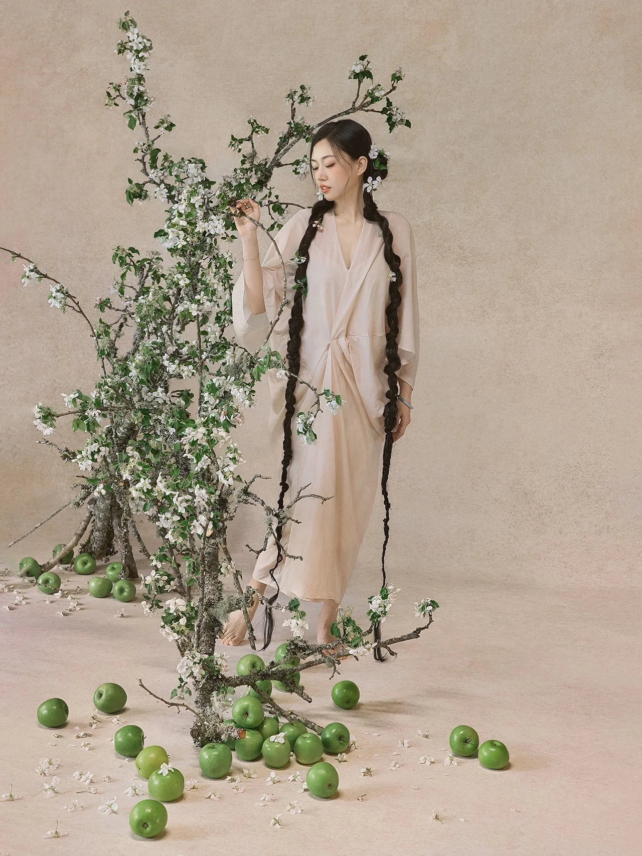 A woman dressed in a flowing beige gown stands next to a blossoming tree with white flowers. Her long, dark hair is styled in two thick braids adorned with small white flowers. Green apples are scattered on the ground and some are clustered at the ba