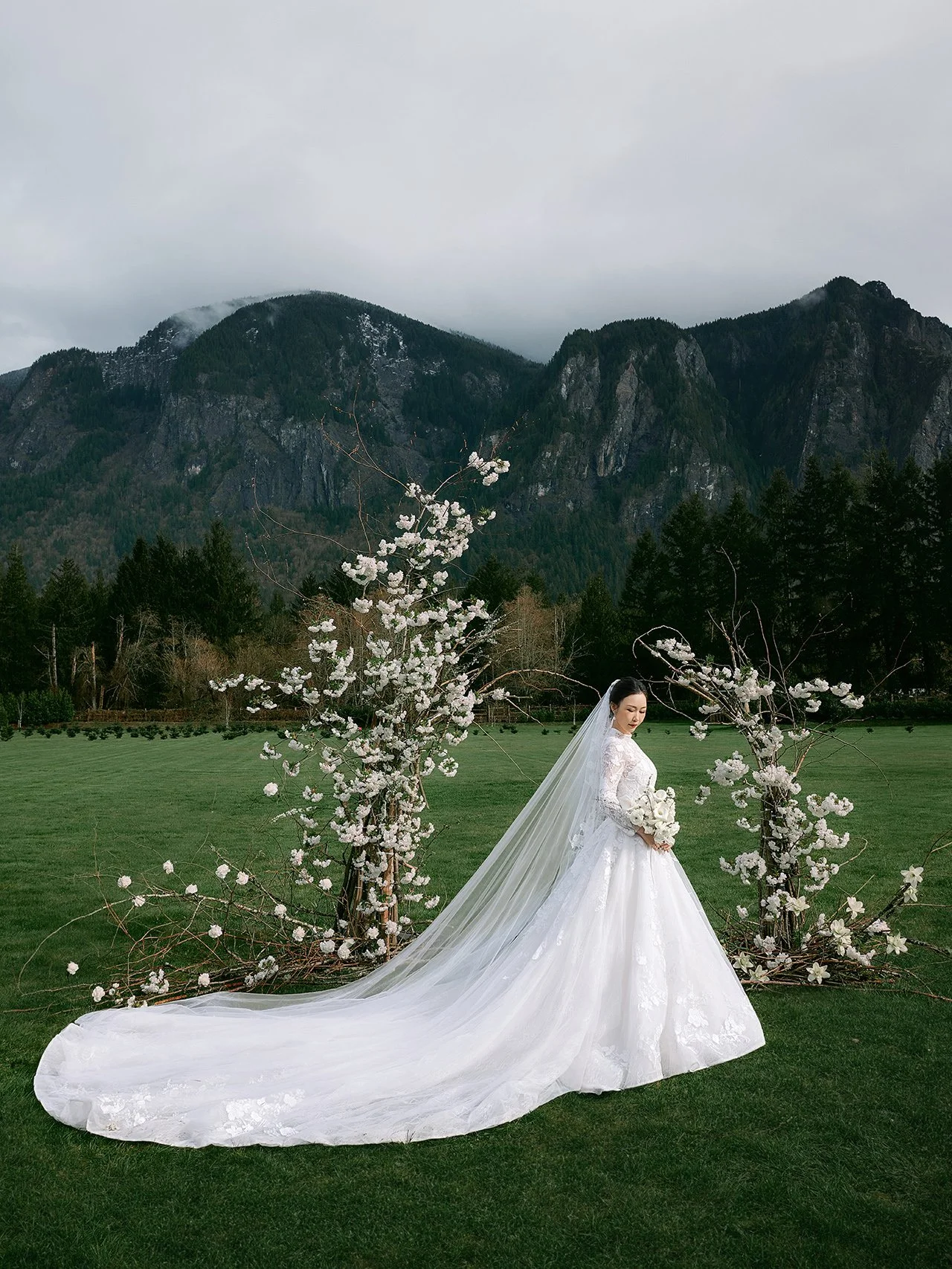A bride in a white wedding gown with long train and veil, standing outdoors with a mountain backdrop. She holds a bouquet of white flowers, and there are flowering branches in front of her.