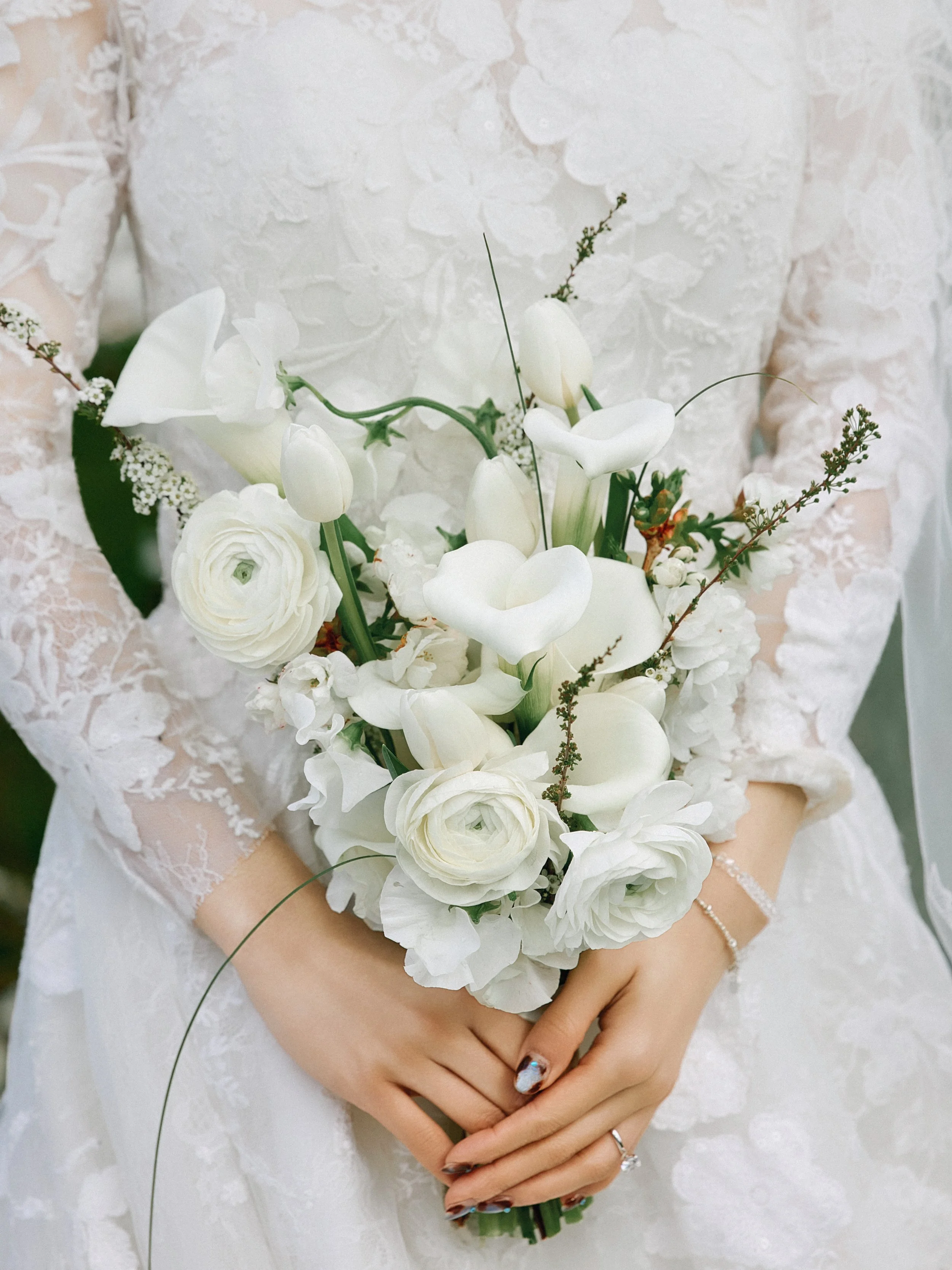 A bride in a lace wedding dress holding a bouquet of white flowers, including white calla lilies and roses, with her hands crossed in front of her waist.