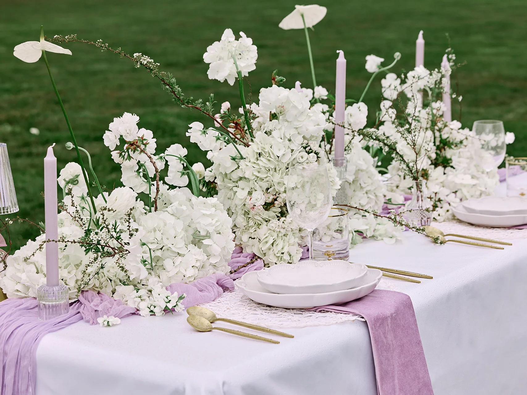 Elegant outdoor table setting with white floral arrangements, lilac table runner, white plates, clear glasses, and tall lilac candles on a green lawn backdrop.