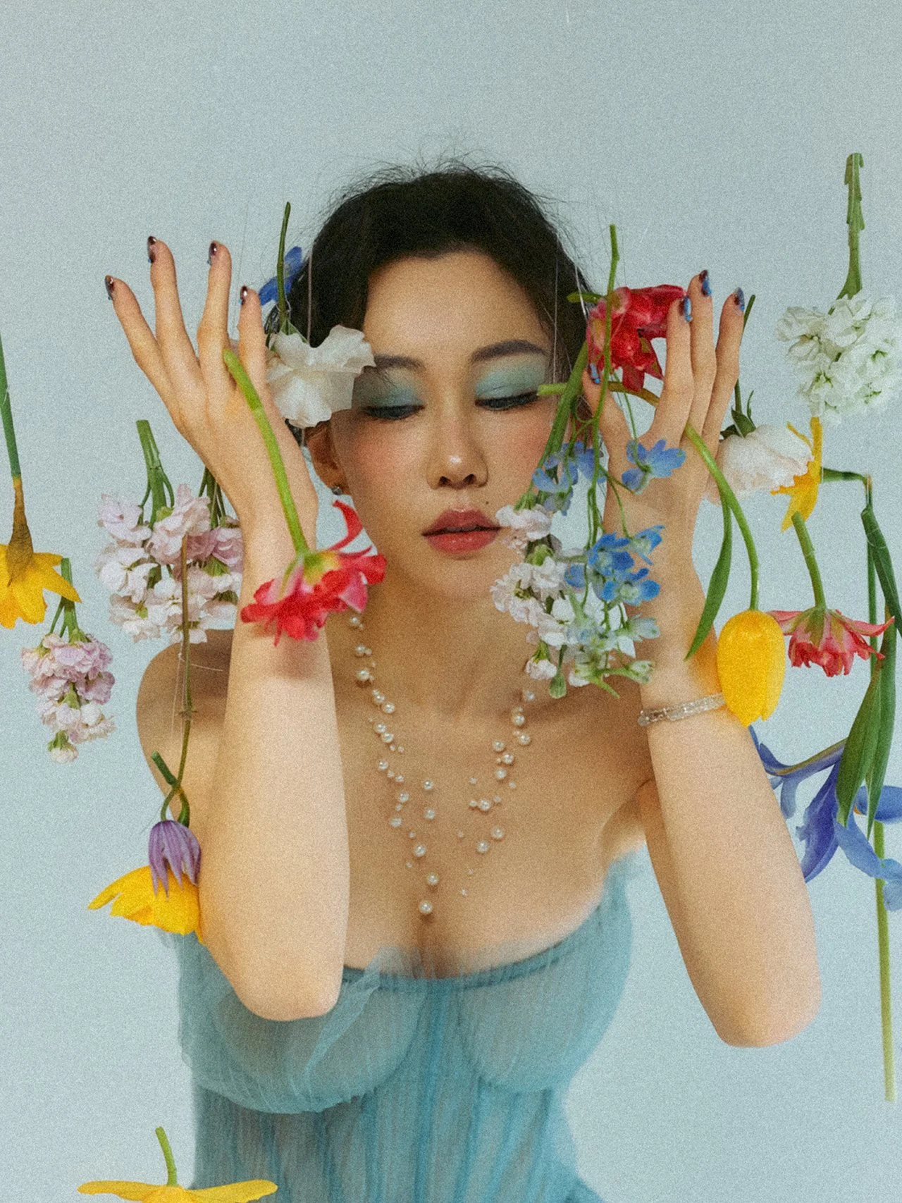 A woman with dark hair and makeup, wearing a pearl necklace and bracelet, surrounded by colorful flowers with her eyes closed and hands near her face.