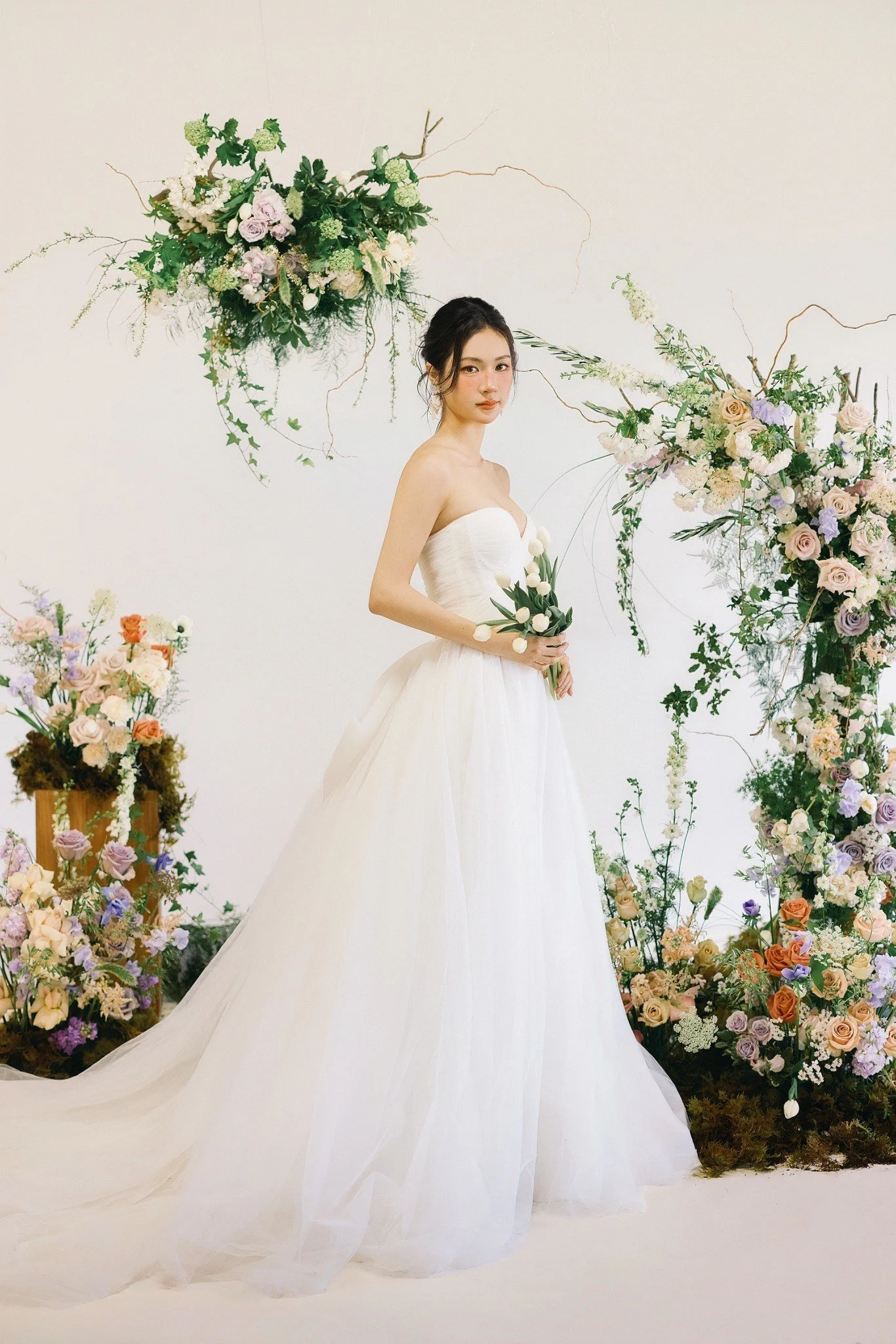 A woman in a white wedding dress holding a bouquet of white tulips, surrounded by large floral arrangements of pastel roses, hydrangeas, and greenery on a white background.
