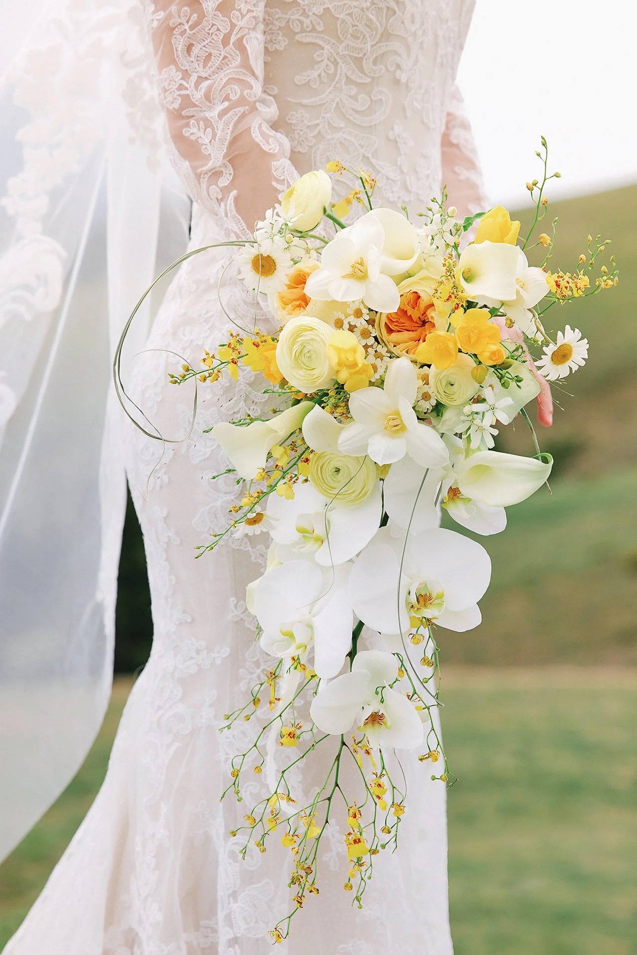 Bride holding a cascading bouquet of white and yellow flowers, including orchids, roses, and daisies, with intricate lace on her dress.