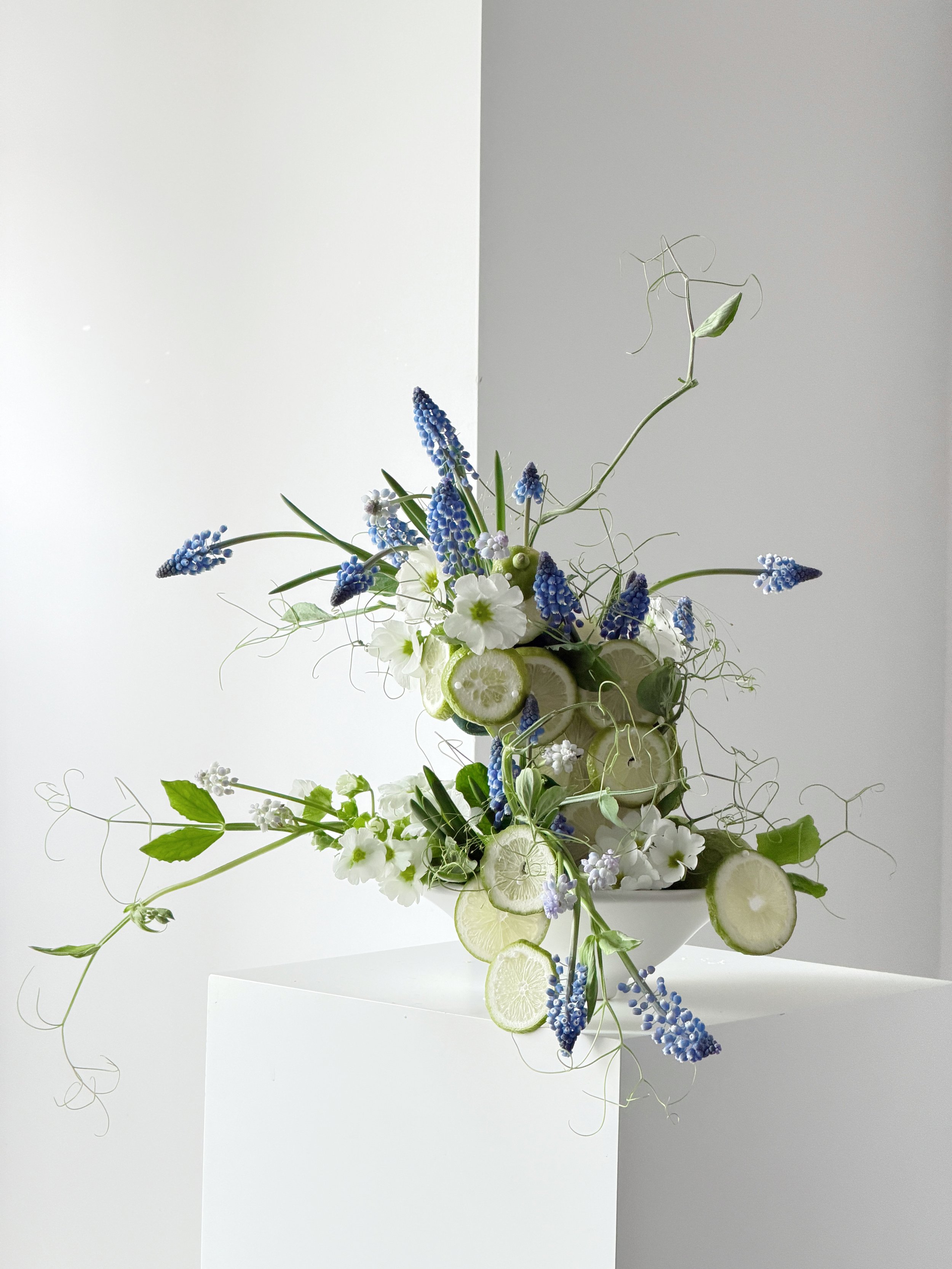 A tall, elegant flower arrangement in a white vase with lime slices, white and purple flowers, and green foliage, set against a two-toned gray and white background.