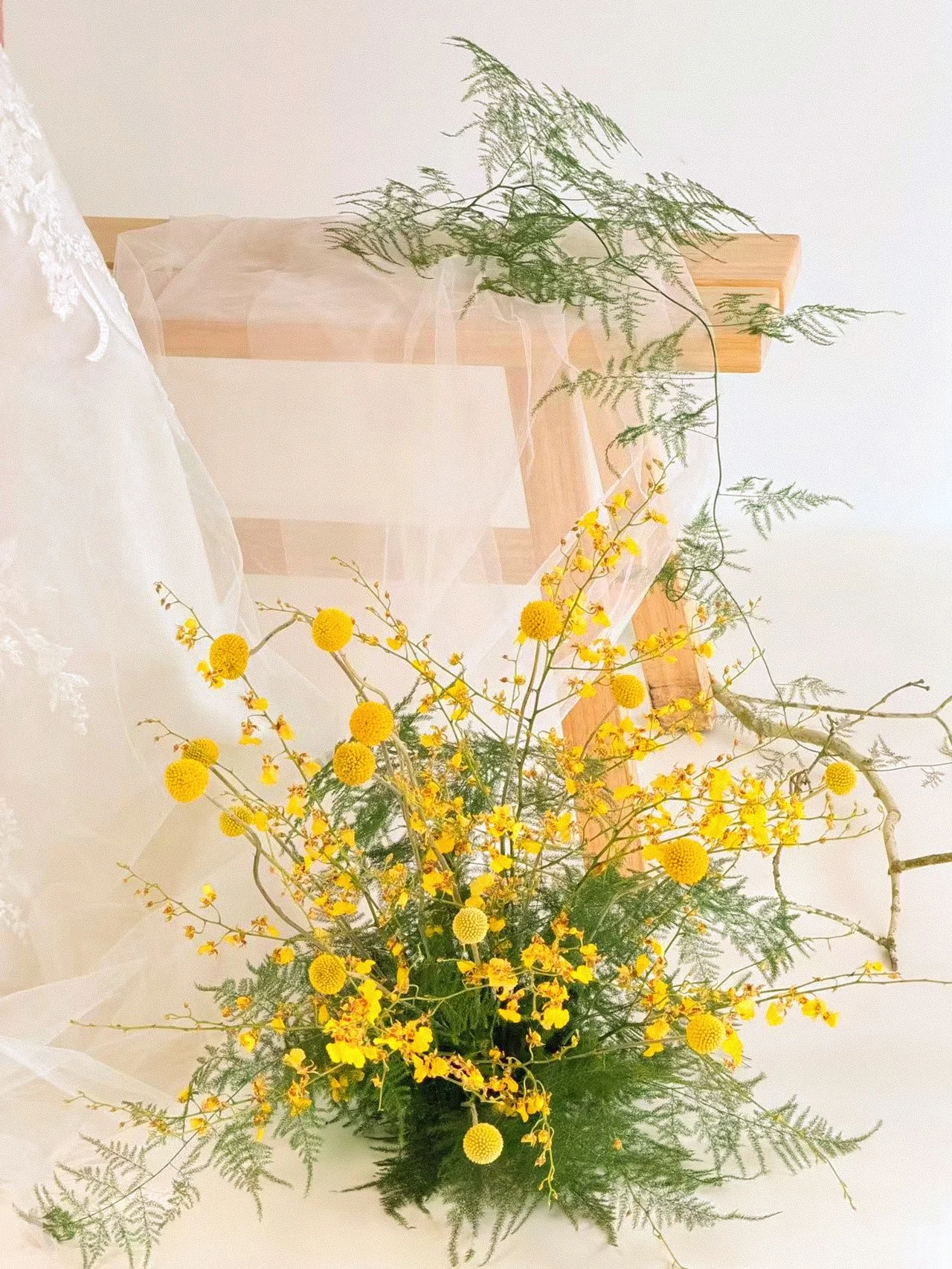 Arrangement of yellow flowers, including craspedia and forsythia, with green fern foliage in a clear glass vase.