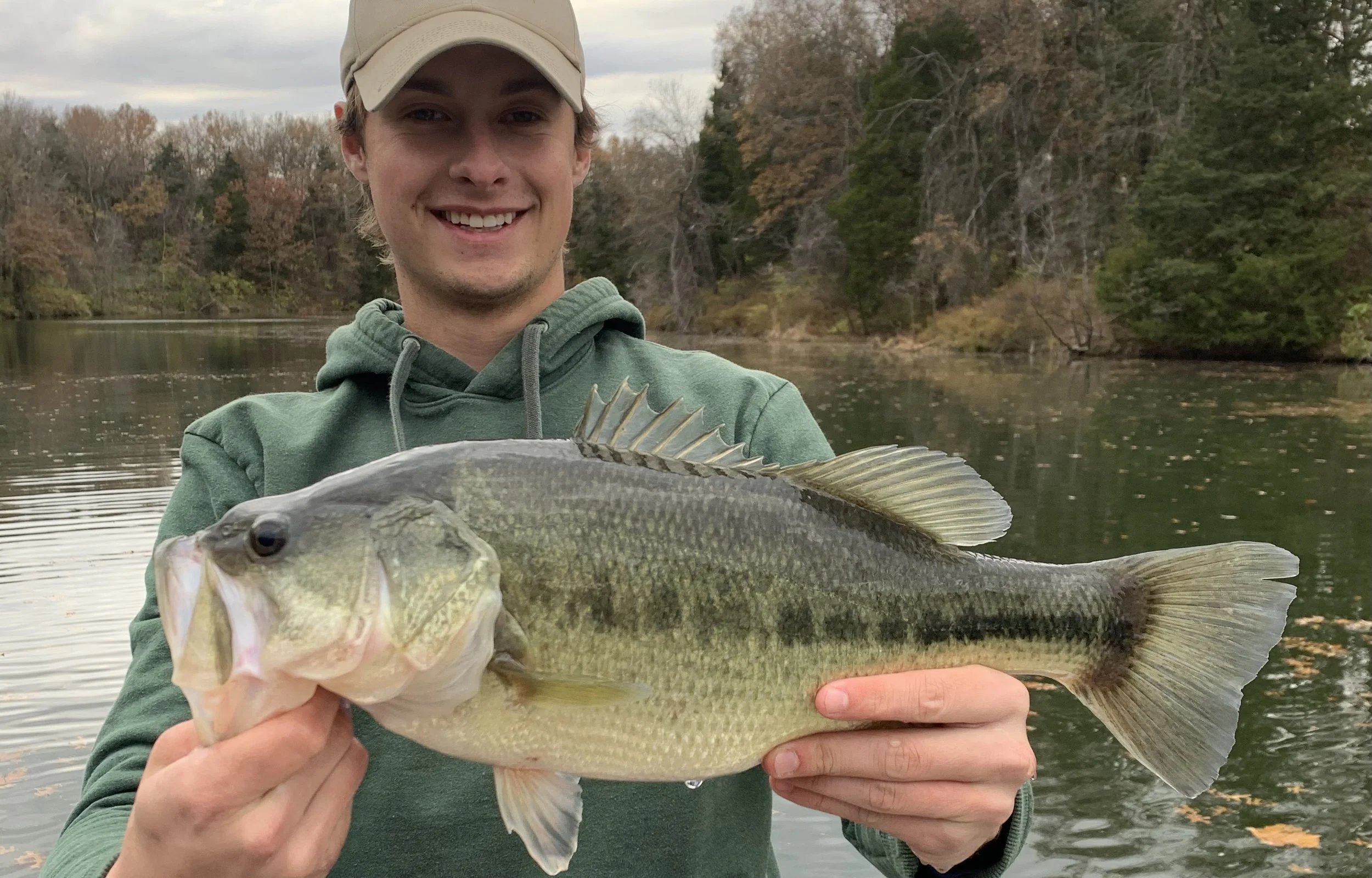 Young man wearing a beige cap and green hoodie holding a large fish, smiling by a calm lake with trees in the background.