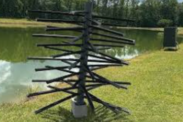 An outdoor sculpture resembling a Christmas tree made of black metal branches on a grassy area next to a body of water.