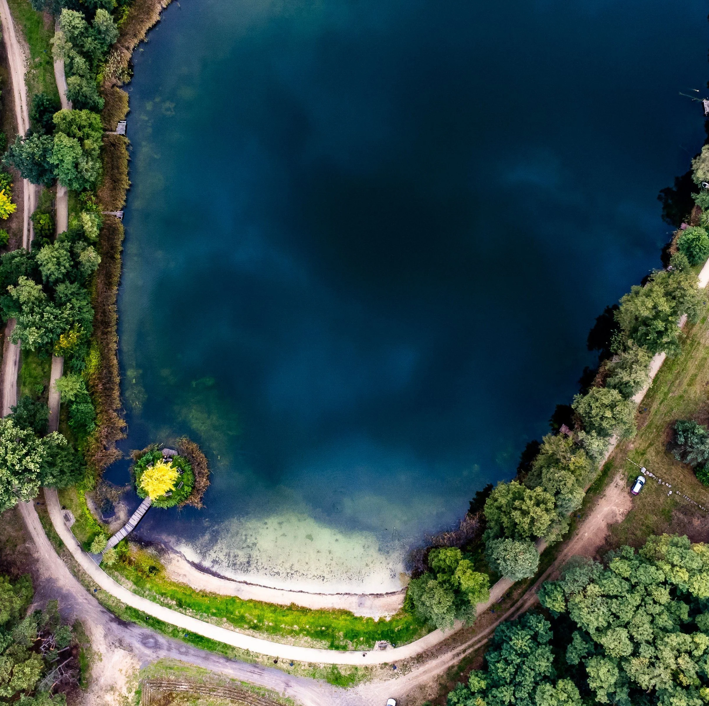 Aerial view of a lake surrounded by trees and a winding dirt path with parked cars.