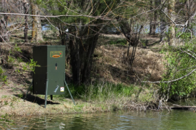 A green electrical box near a riverbank with trees and grass in the background.