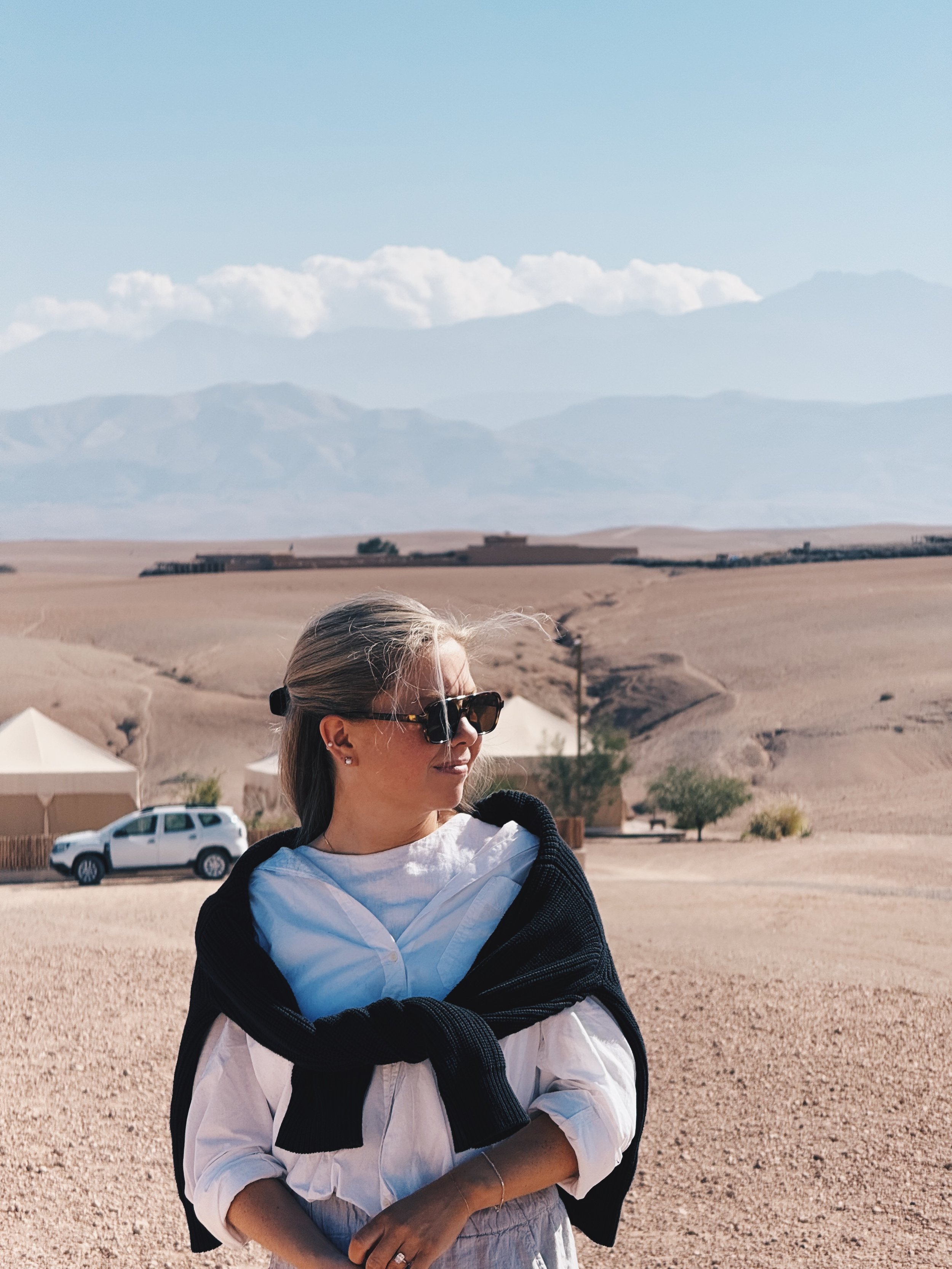 A woman with sunglasses and a white shirt, with a black sweater tied around her shoulders, standing in a desert landscape with mountains in the background.